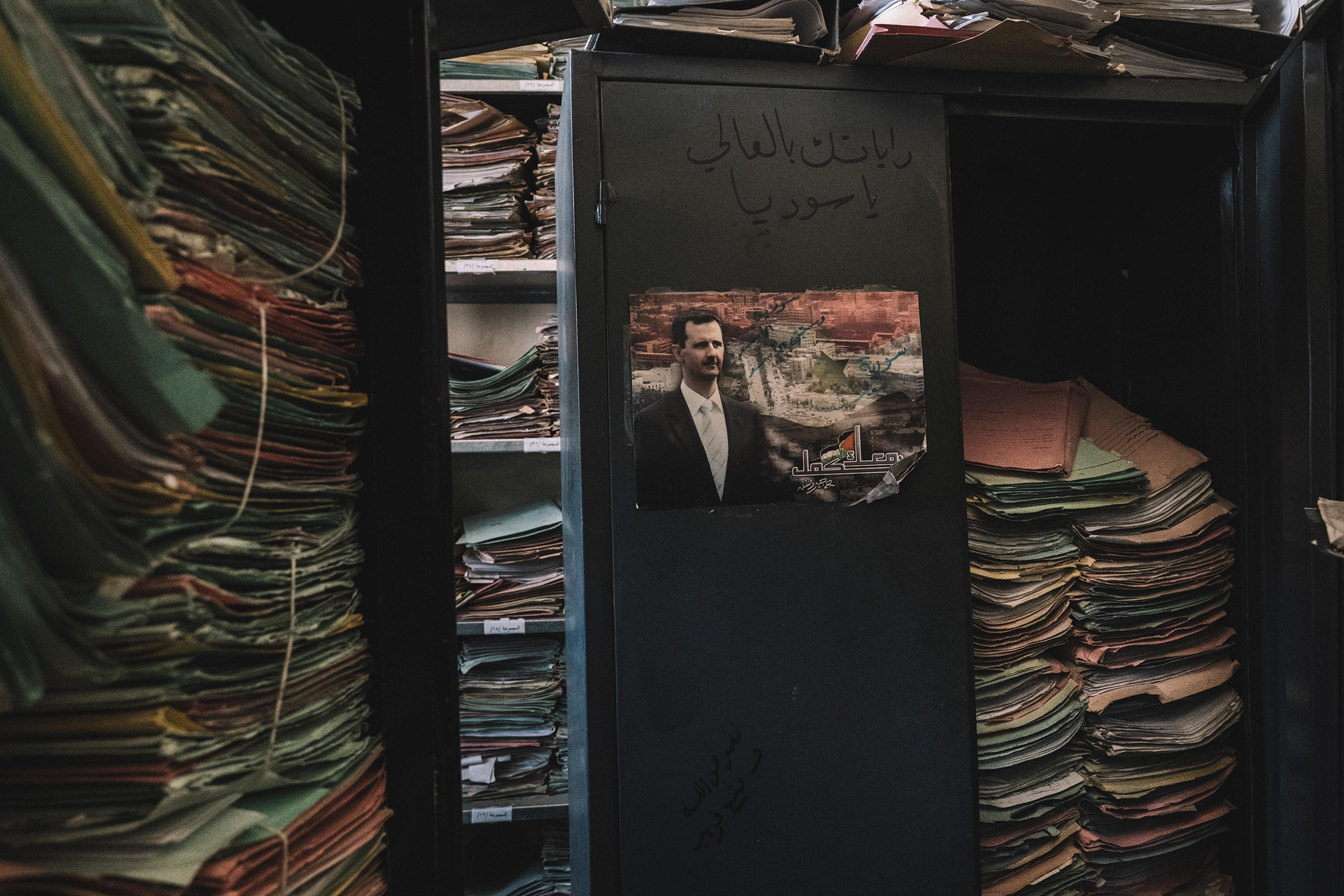 A cluttered archive with stacks of files in cabinets, a poster on the cabinet door, and dark ambient lighting.