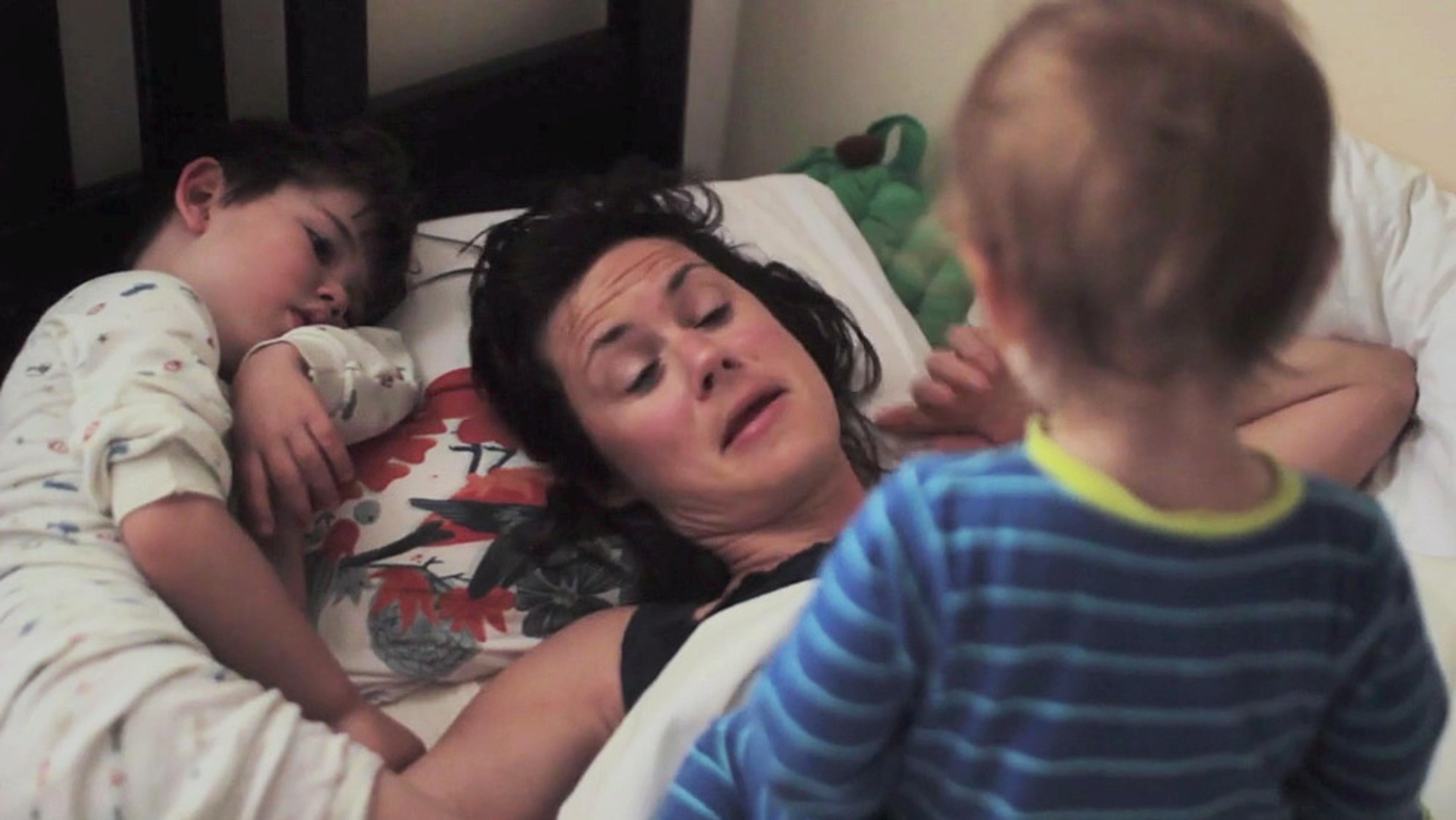 A tired woman lying in bed with a child in pyjamas next to her and a toddler in striped pyjamas standing near the bed.