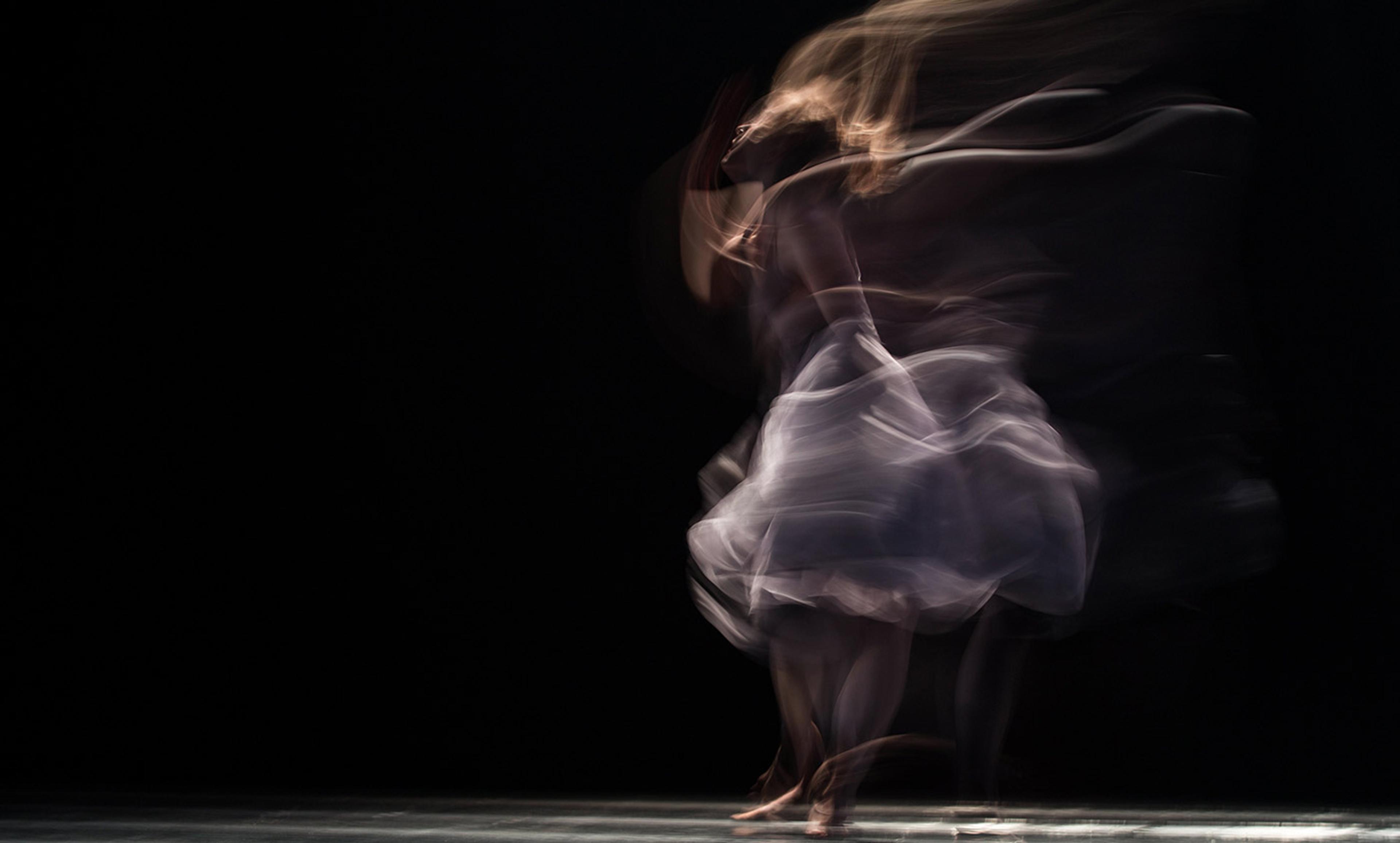 A dancer in motion on a dark stage. The blurred effect creates a sense of fluid movement and ethereal light.