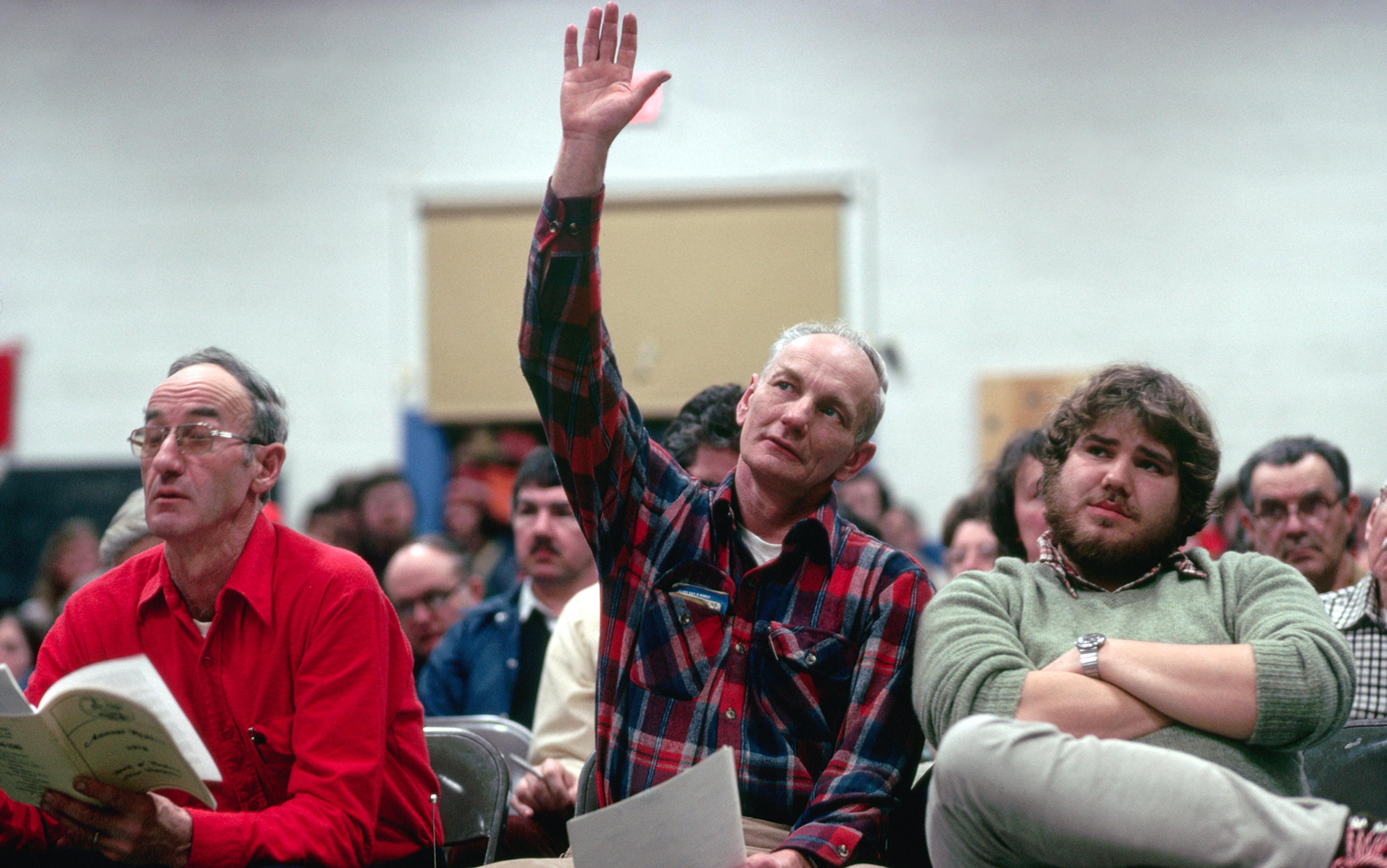 Photo of three men seated among others, one in the centre raising his hand, another reading a booklet, all engaged in an indoor event.