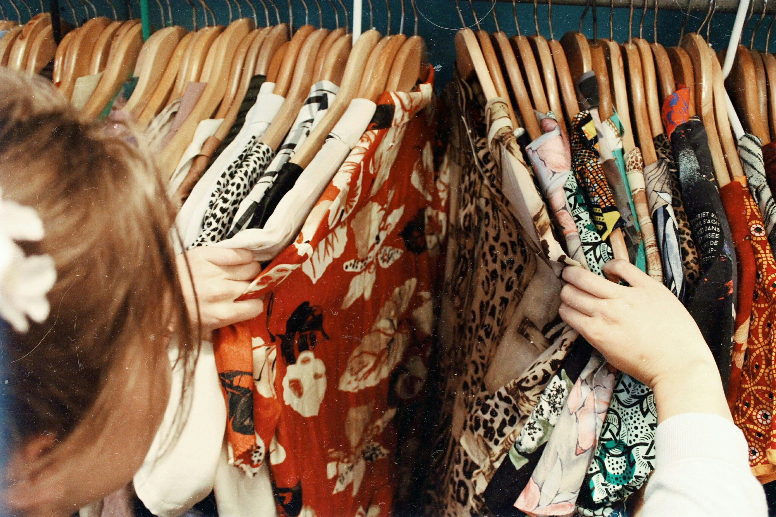 Photo of a person browsing colourful clothes on wooden hangers in a wardrobe, highlighting diverse patterns and textures.
