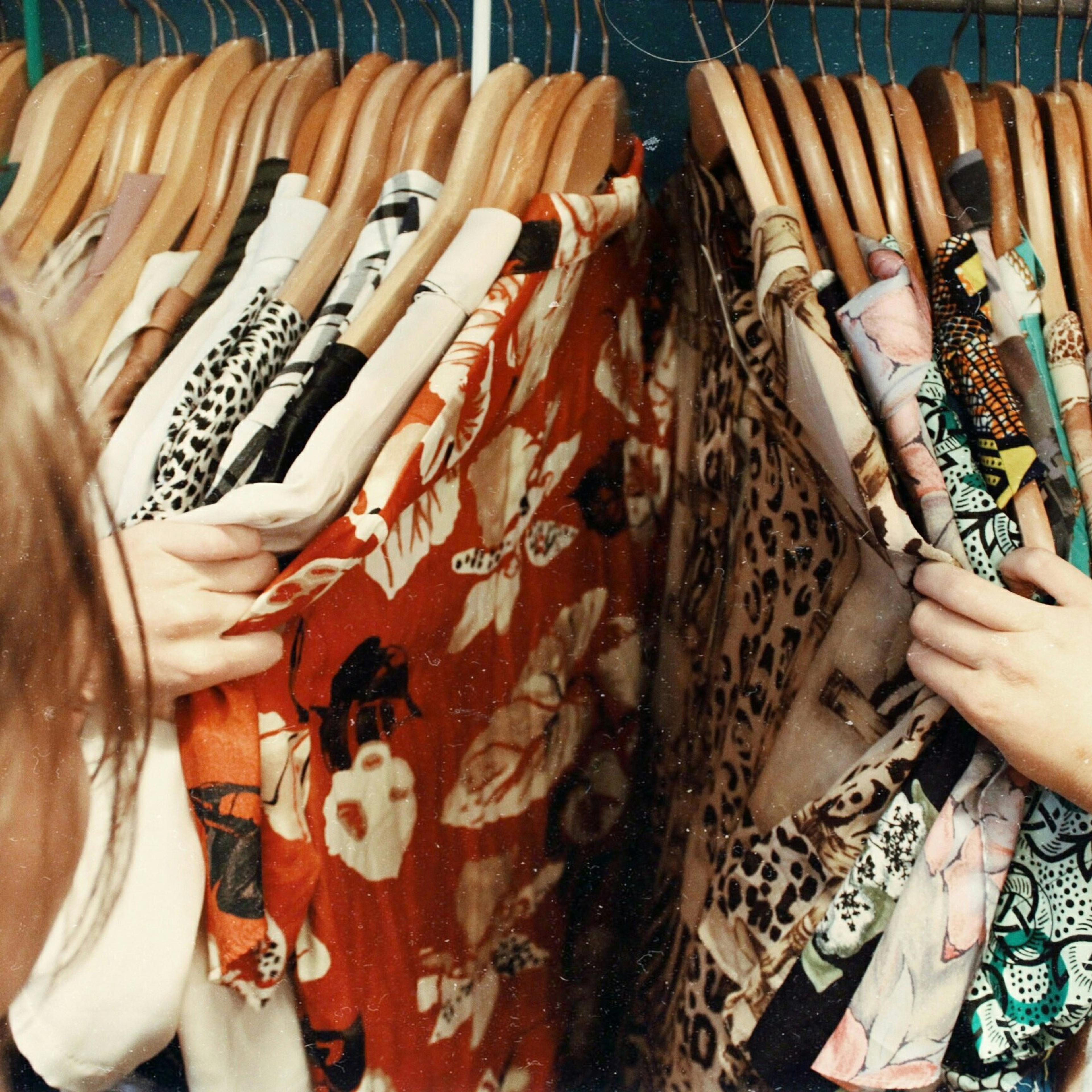 Photo of a person browsing colourful clothes on wooden hangers in a wardrobe, highlighting diverse patterns and textures.