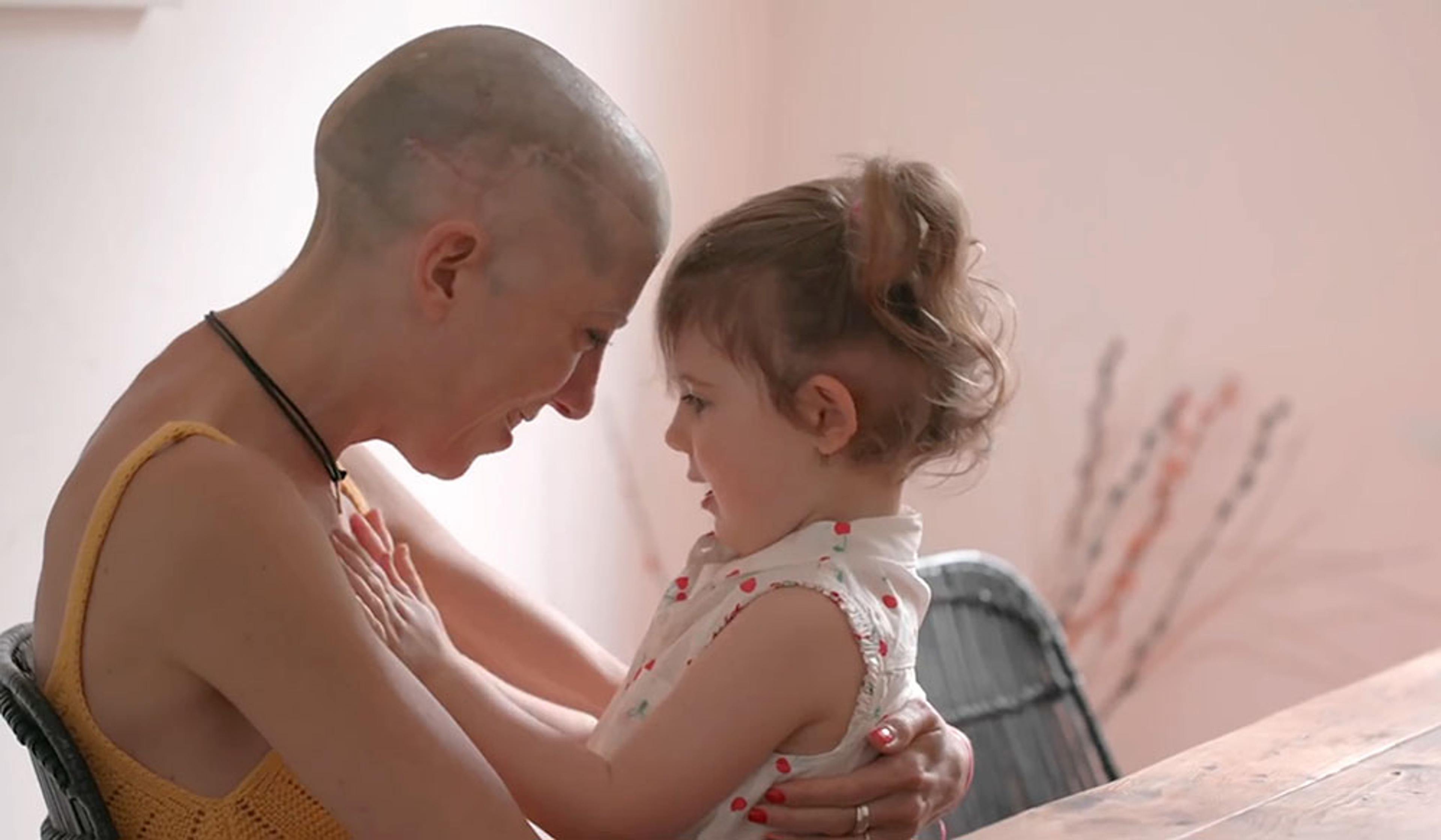 A woman with a shaved head holds young child in her lap. Both are smiling, touching foreheads and embracing.