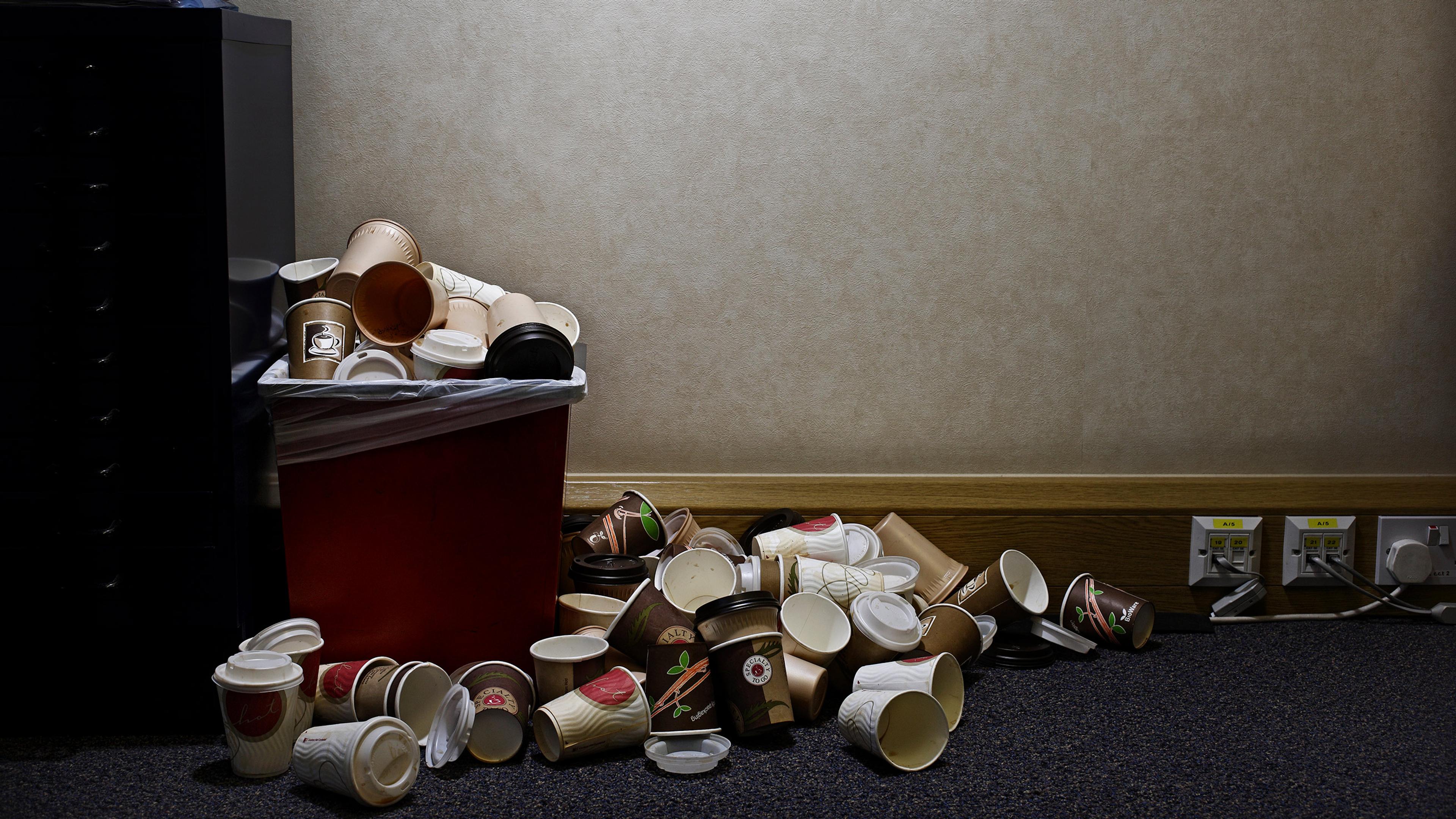 A red bin overflowing with paper coffee cups next to a wall with powerpoints in an office setting.