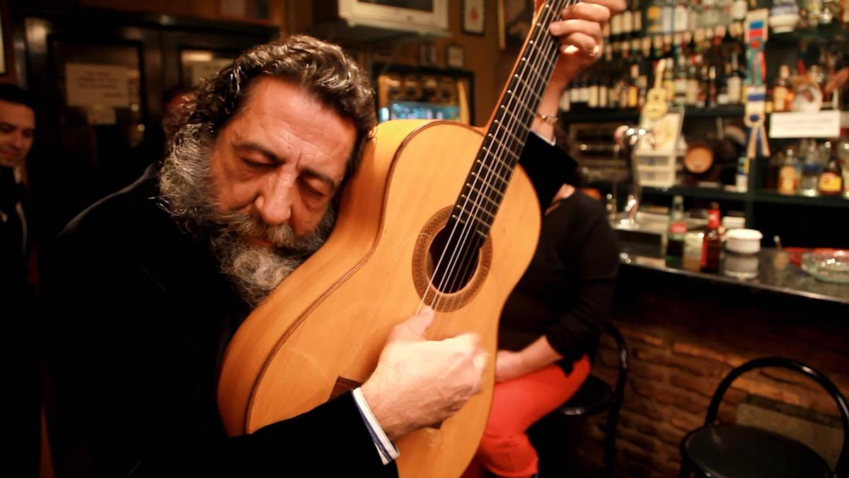 Photo of a bearded man embracing a guitar in a cosy bar setting, eyes closed, surrounded by bottles and seated patrons.