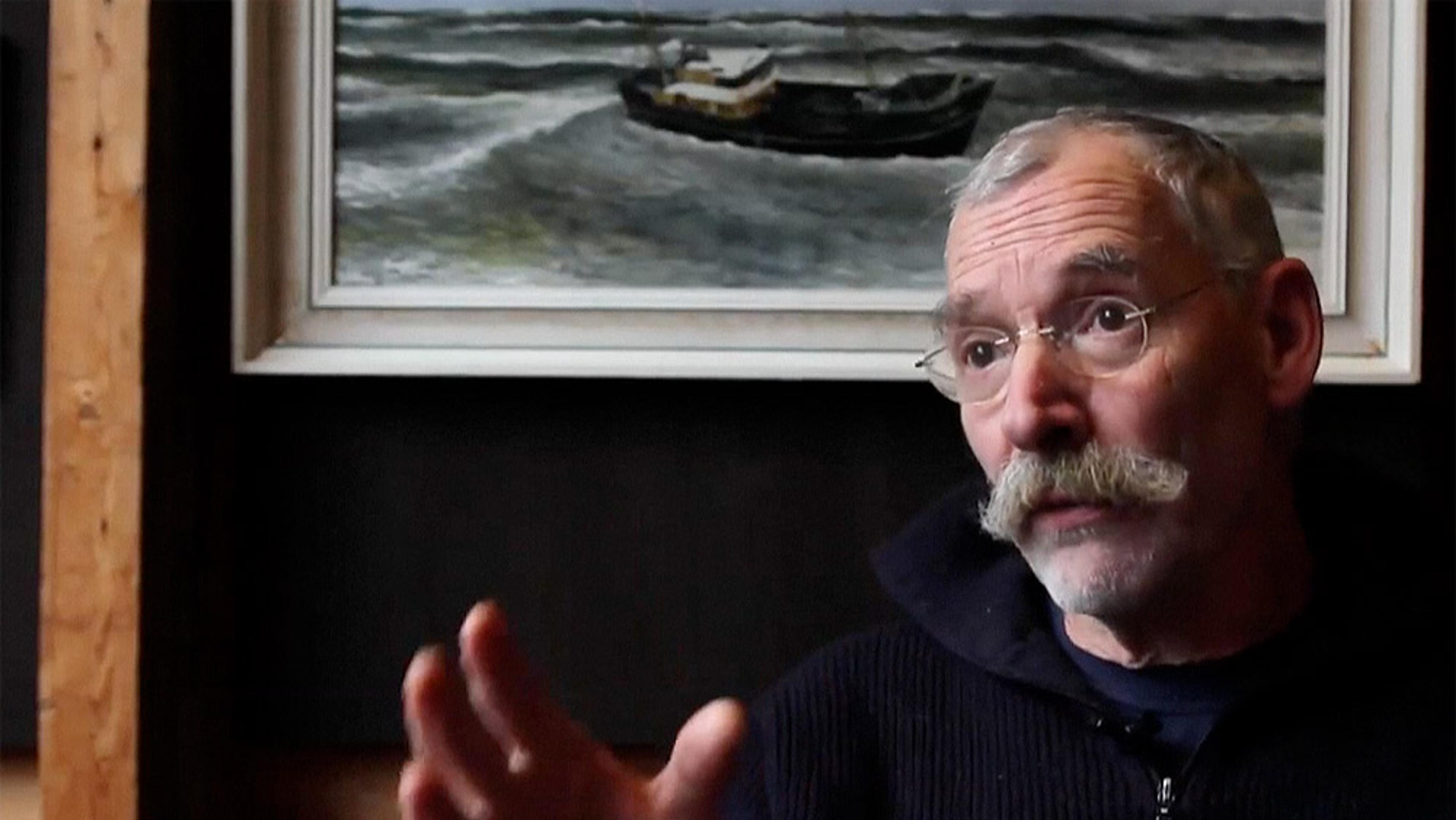 A man with a moustache and glasses gesturing with his hand, a maritime painting of a boat on rough seas in the background.