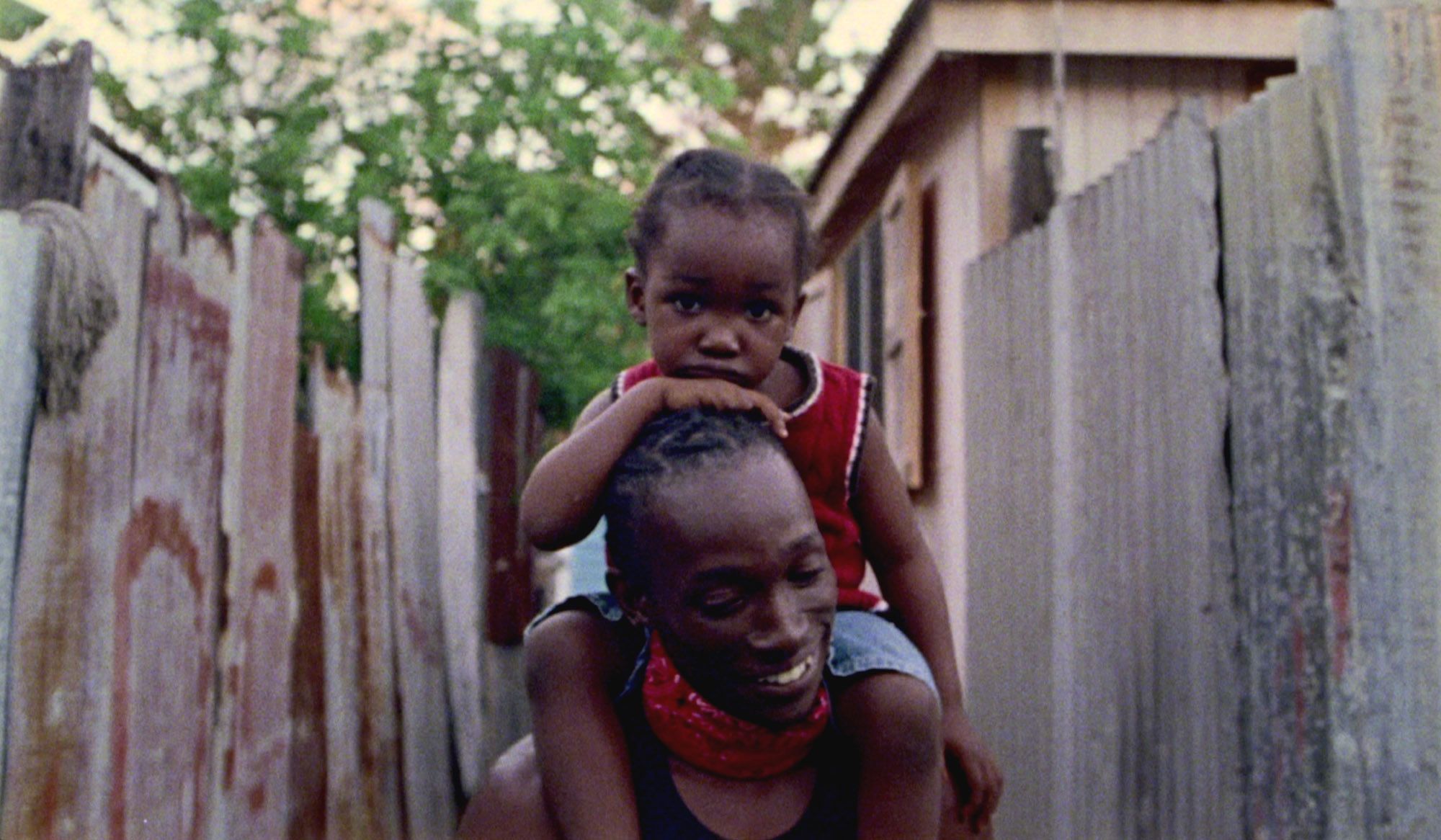 A smiling man carrying a child on his shoulders in an alley with corrugated metal fences and trees in the background.
