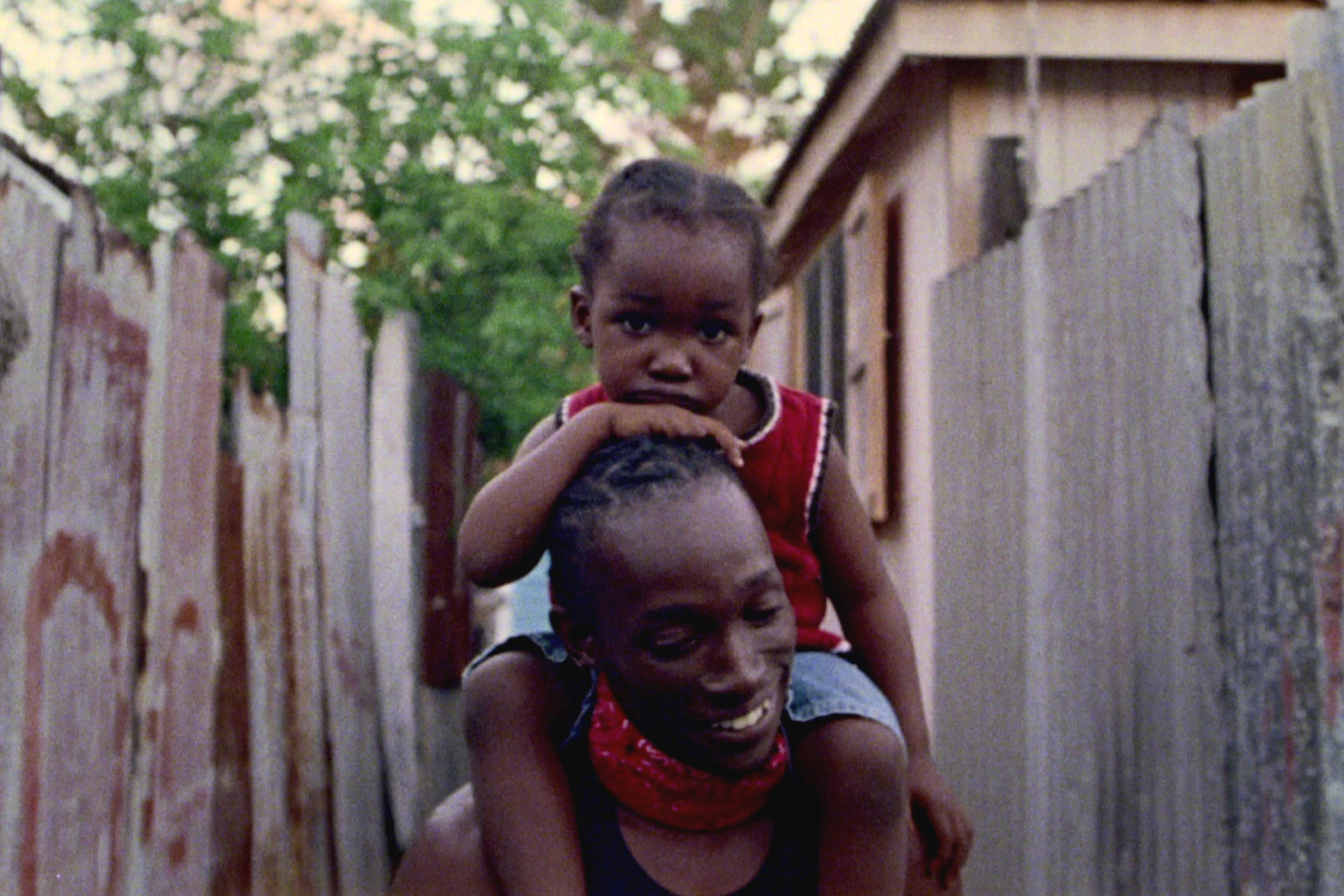 A smiling man carrying a child on his shoulders in an alley with corrugated metal fences and trees in the background.