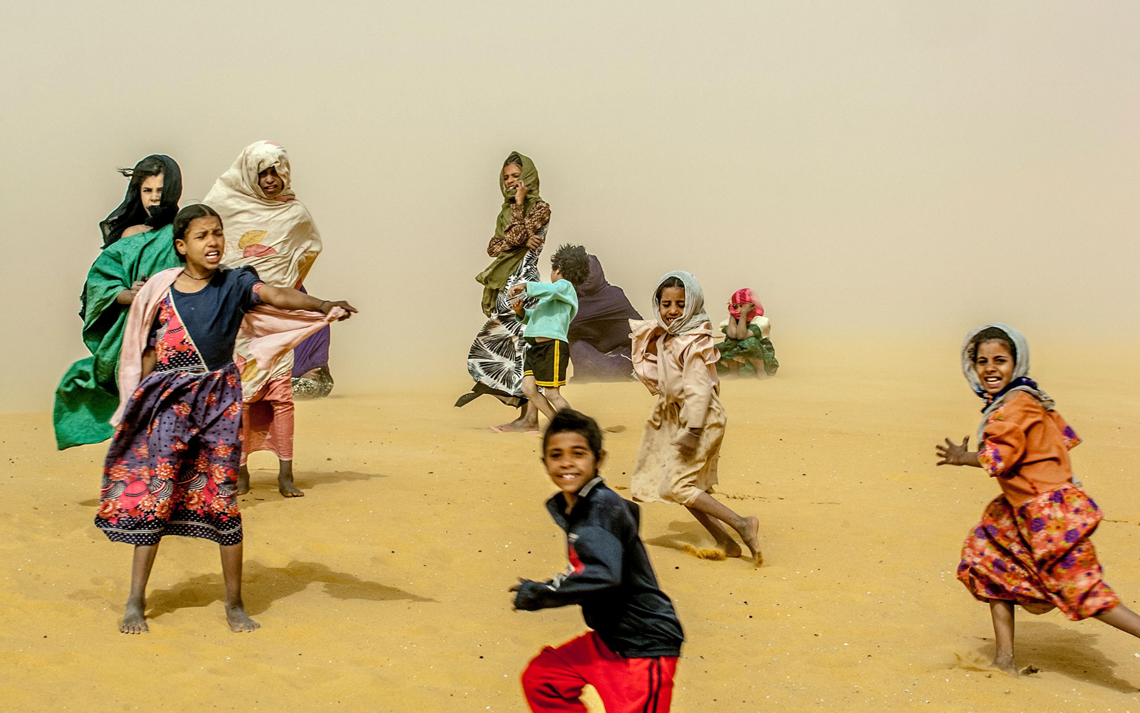 Photo of children playing in the desert in windy conditions, with sand swirling around them and smiles on their faces.