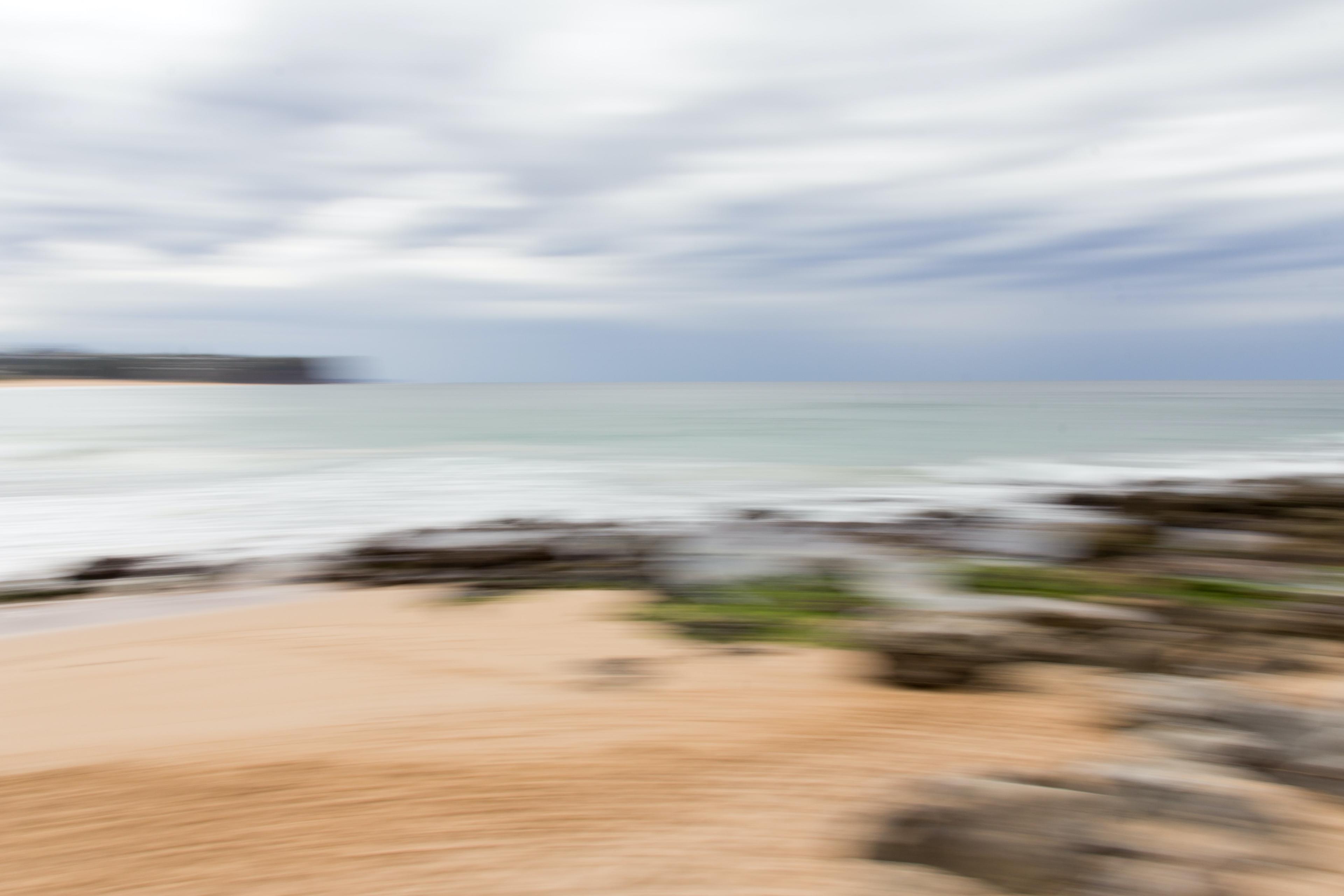 Blurry photo of a coastal scene with sandy beach rocks ocean and cloudy sky conveying a sense of motion or abstraction.