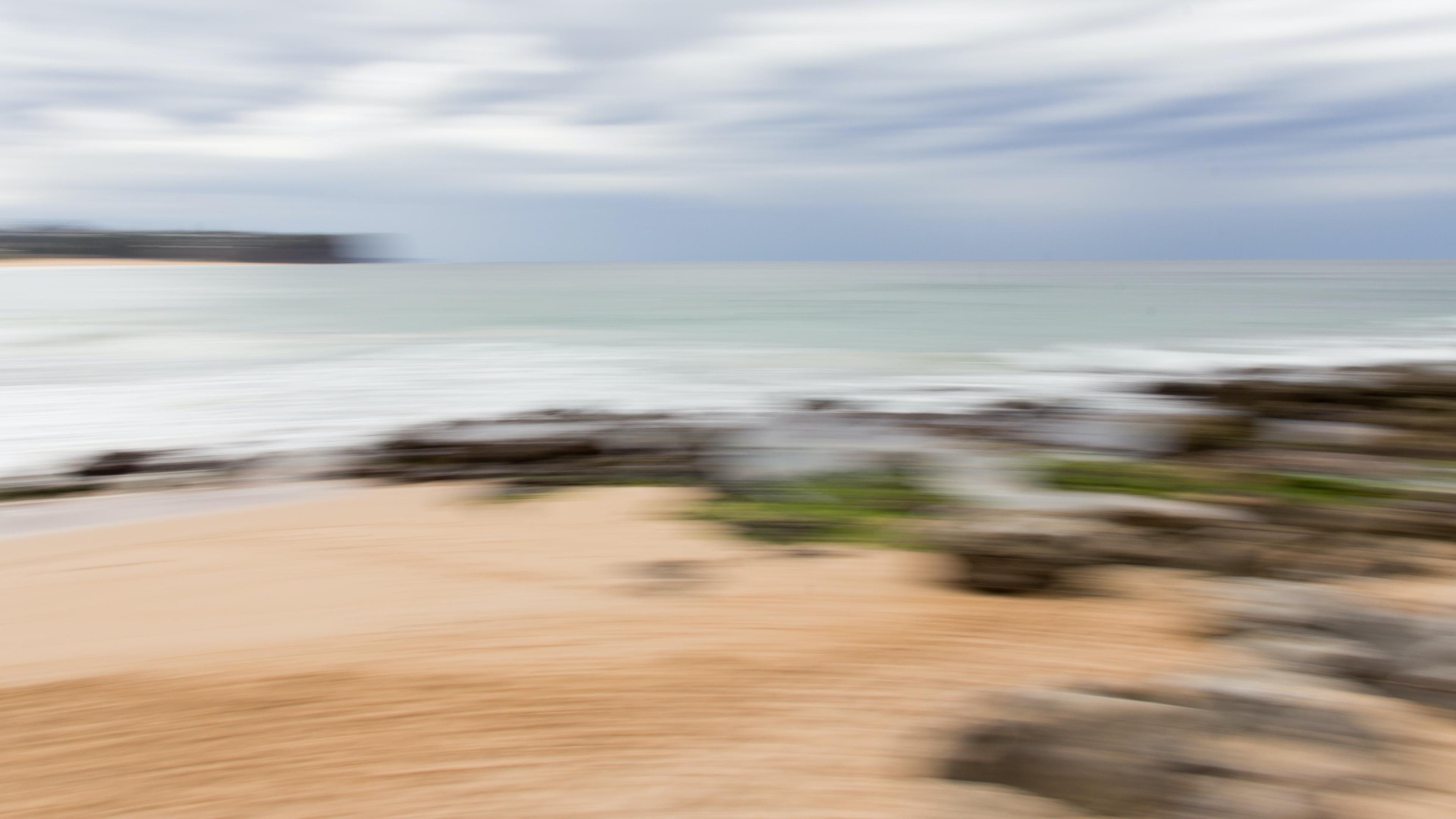 Blurry photo of a coastal scene with sandy beach rocks ocean and cloudy sky conveying a sense of motion or abstraction.