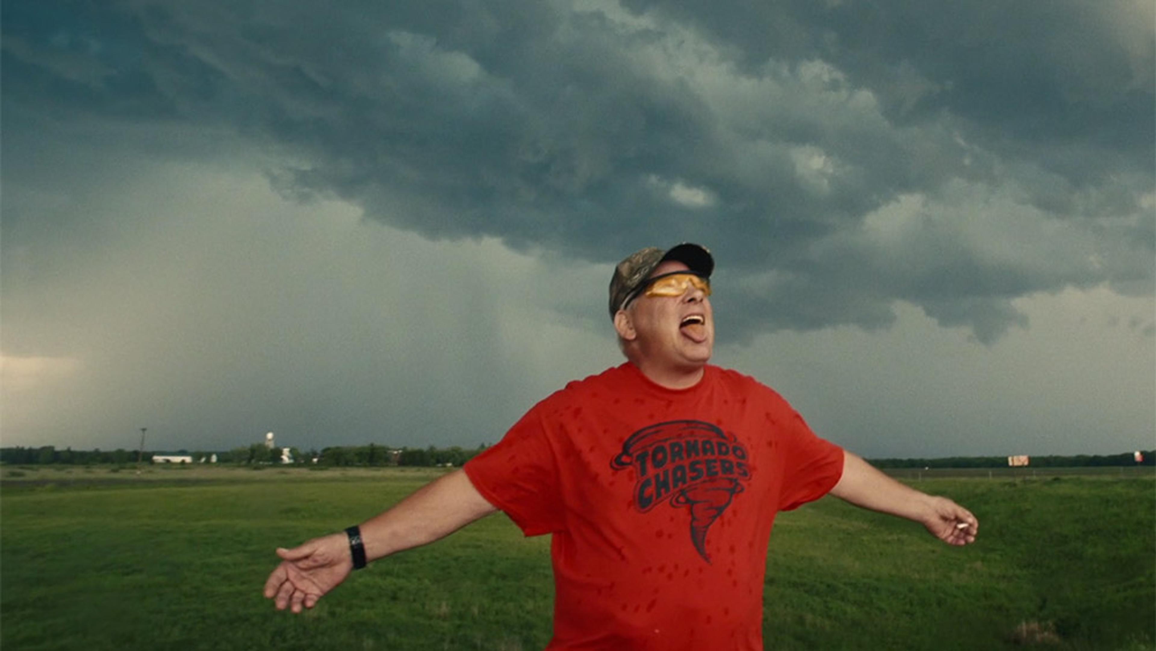 A man in a red “Tornado Chasers” T-shirt standing in a field under a stormy sky with arms outstretched and mouth open.