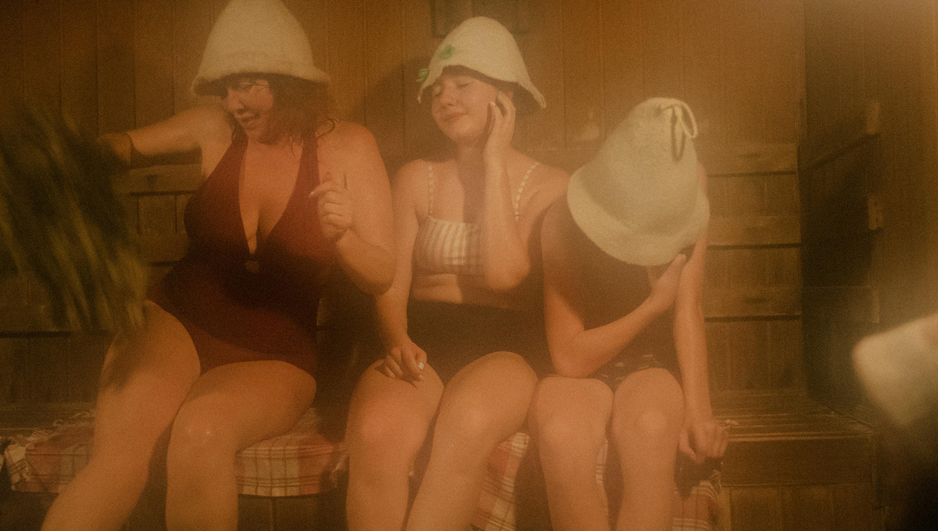 Three people wearing bathing suits and felt hats sit on a wooden bench in a sauna. The sauna is filled with steam, making the ambience hazy. The woman on the left is holding a bundle of twigs, likely for traditional sauna rituals. The mood appears relaxed and warm within the wooden interior.