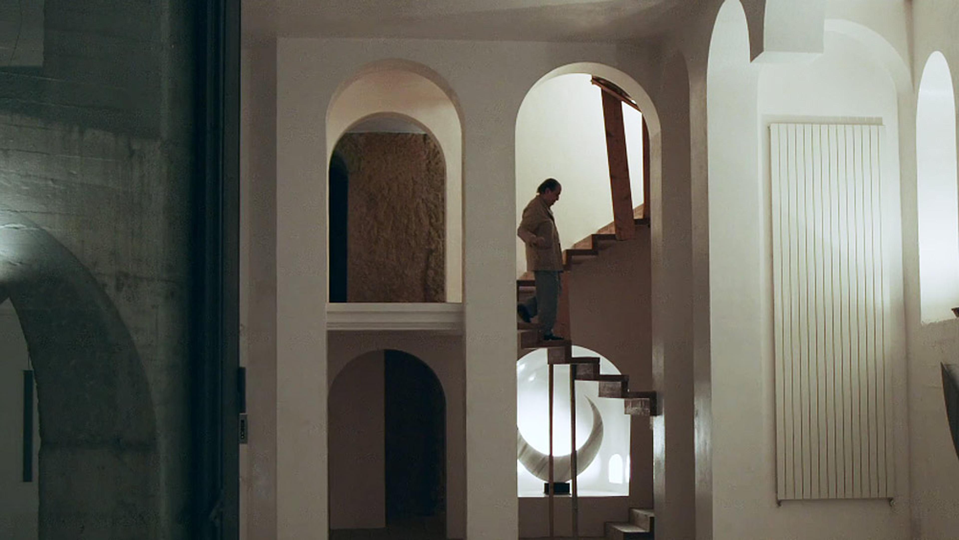 A person descending spiral stairs set within an architectural space featuring arches and minimalist design.
