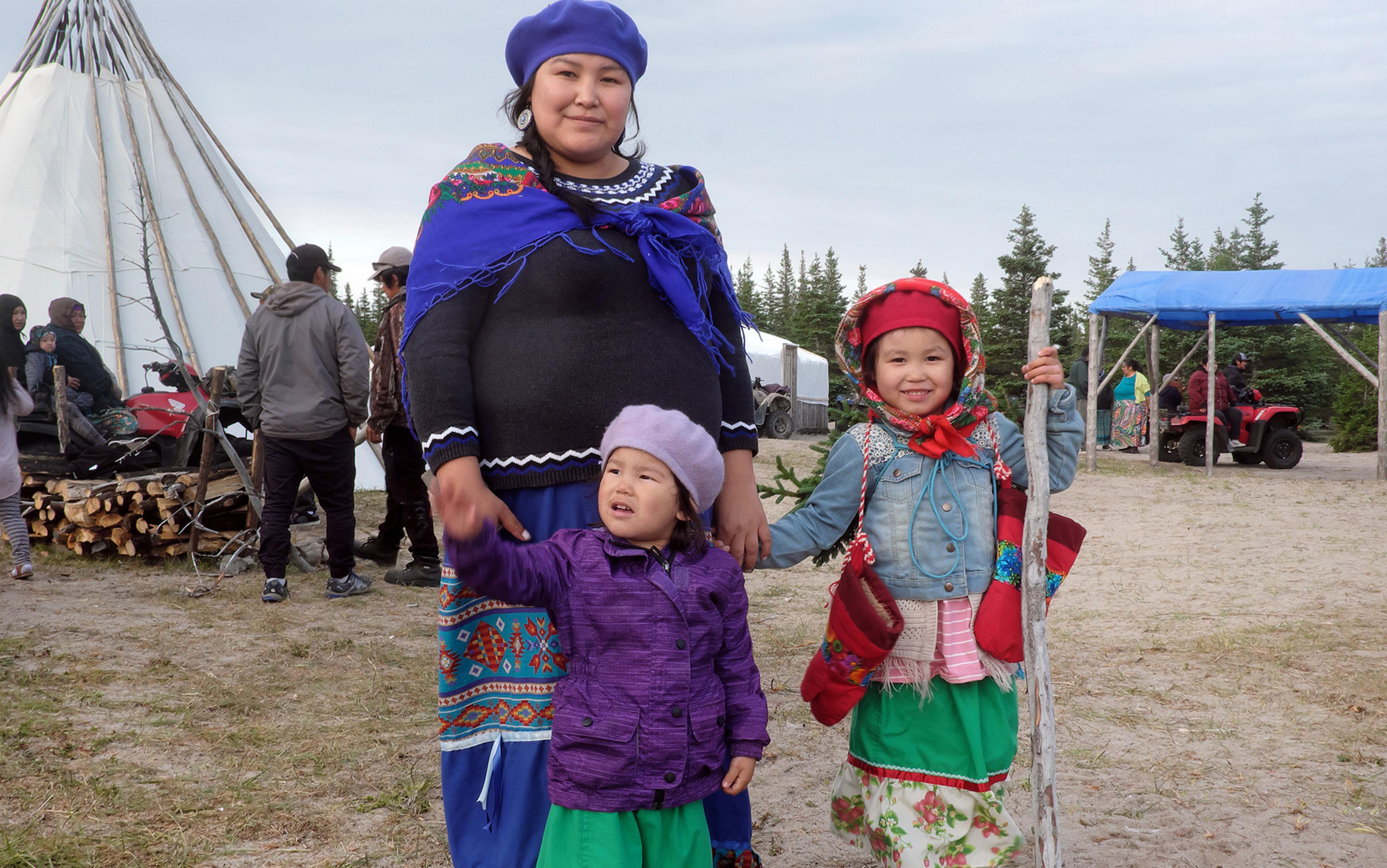 A woman and two children dressed in traditional First Nations clothing stand in front of a teepee structure