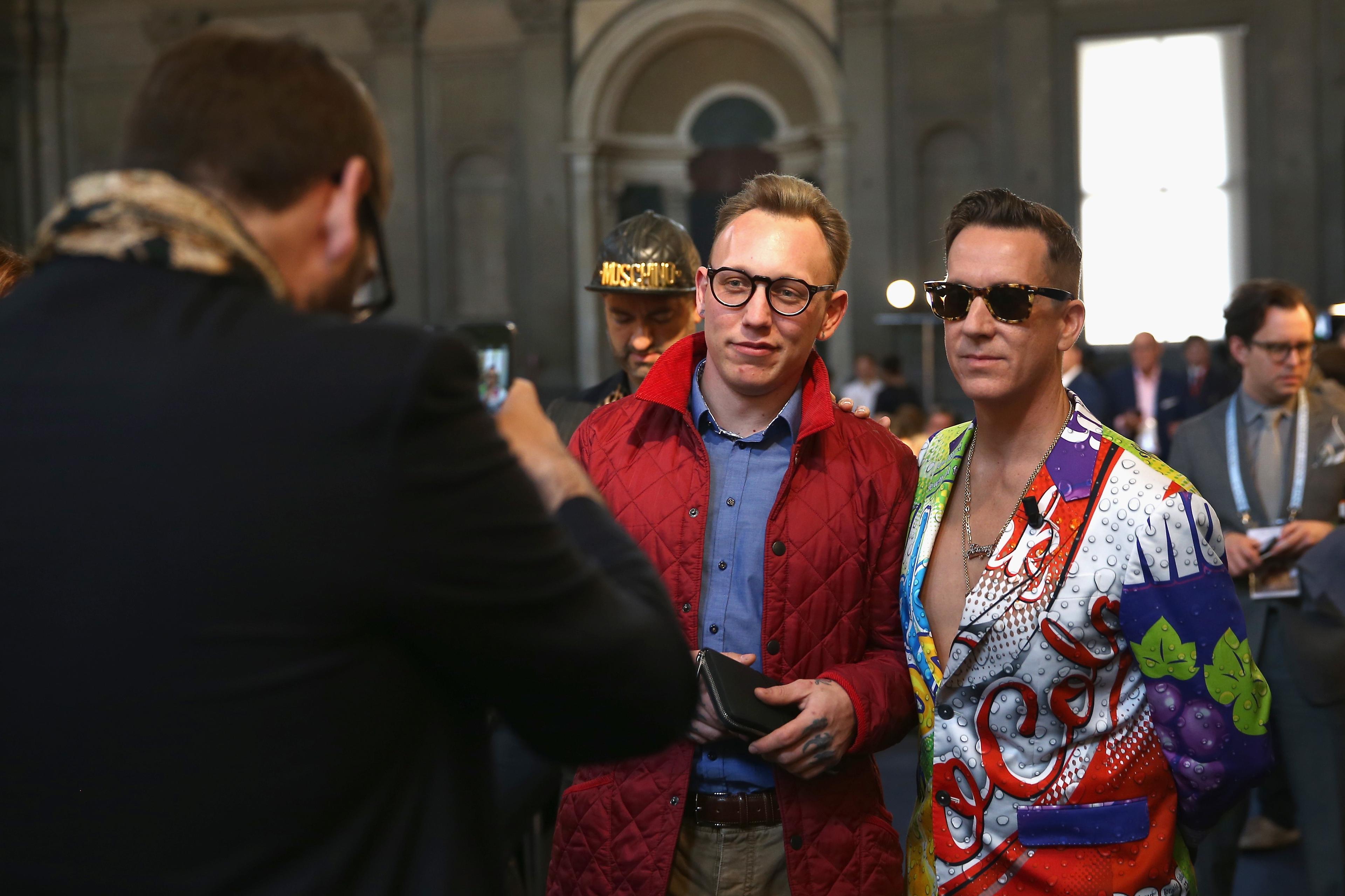 Photo of two men posing for a picture indoors, one in a red coat, the other in a colourful suit with a graphic design.