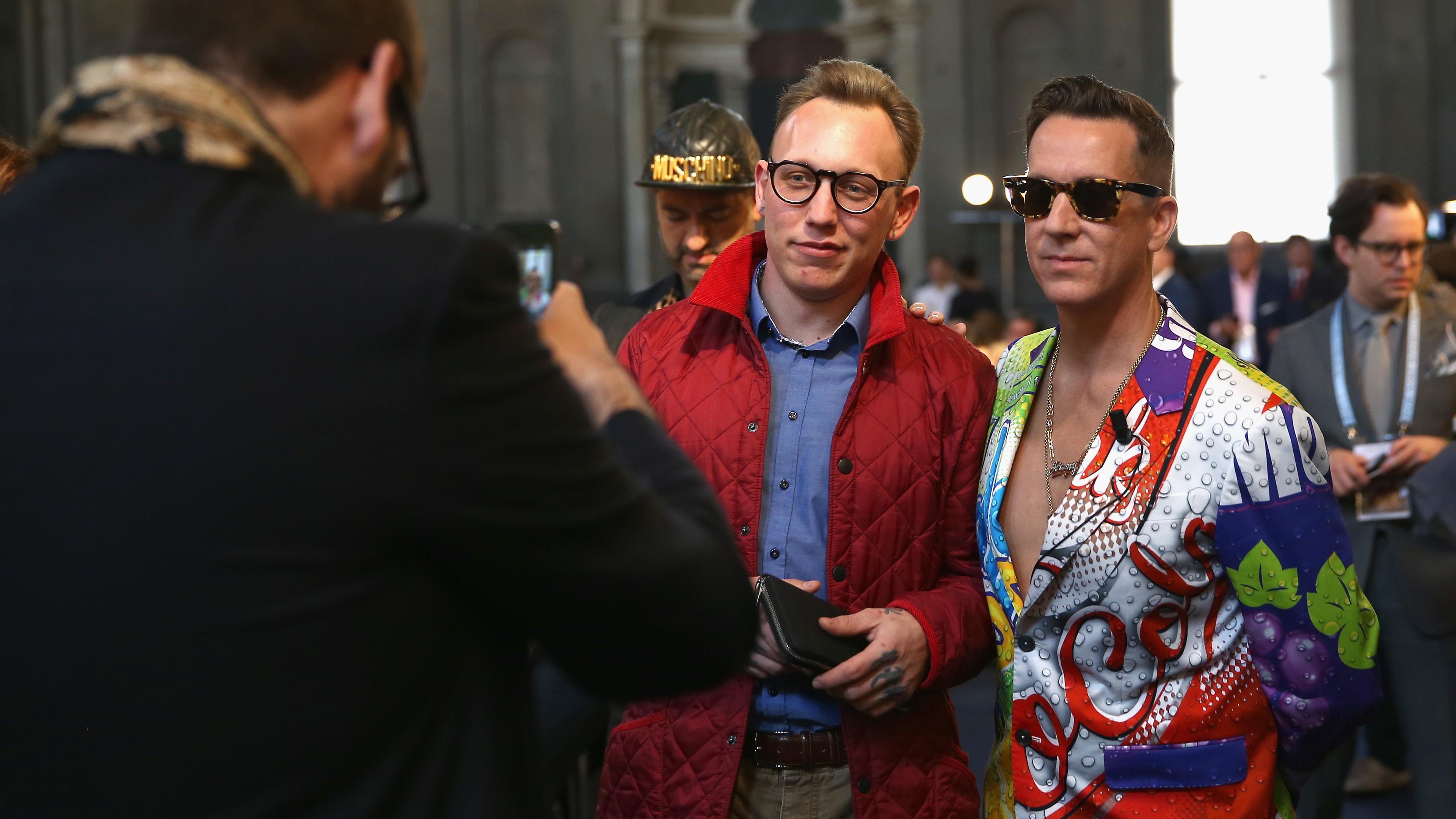Photo of two men posing for a picture indoors, one in a red coat, the other in a colourful suit with a graphic design.