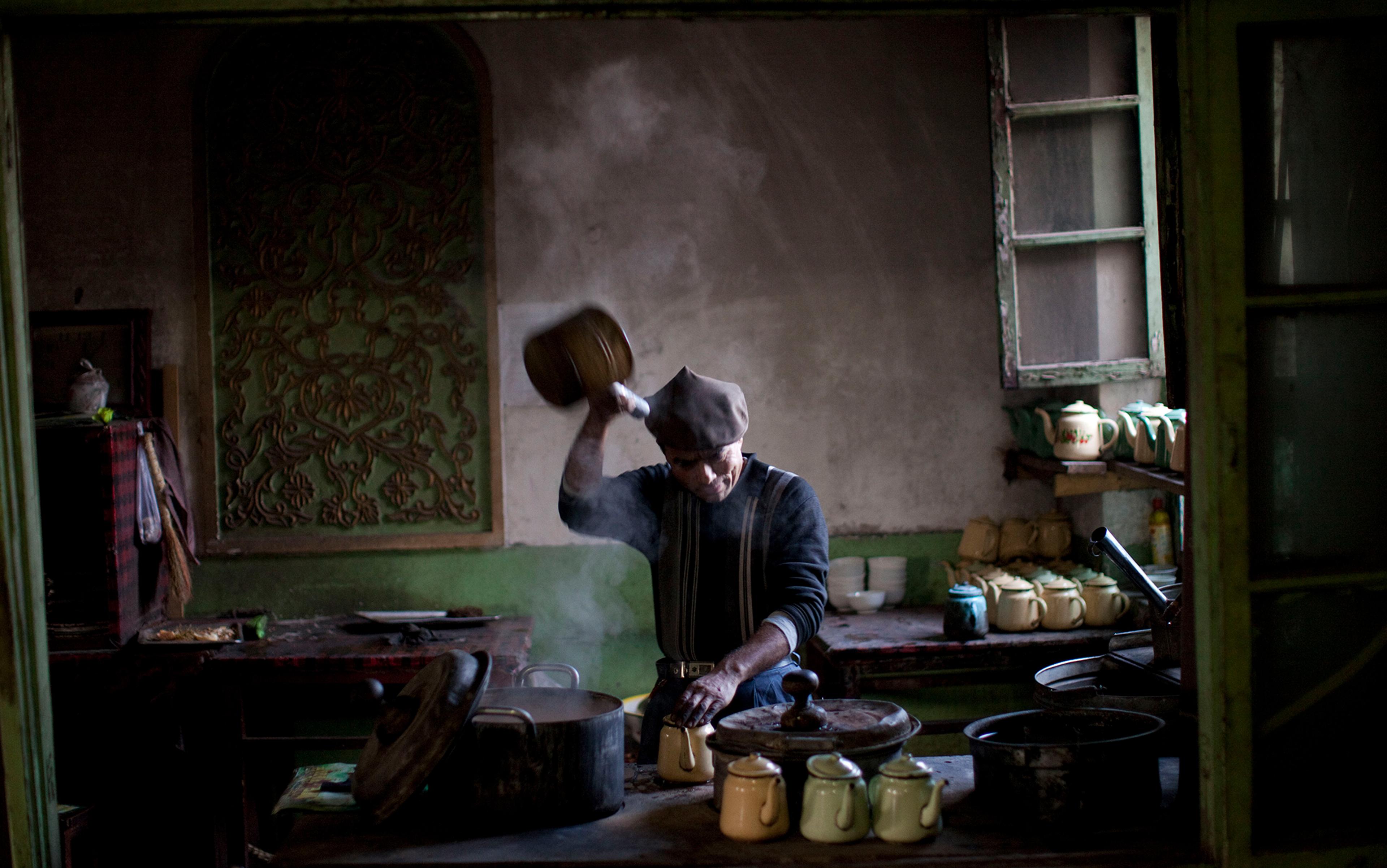 A person in a dimly lit kitchen pouring hot liquid into mugs, with steam rising from the pot, surrounded by teapots and kitchenware.