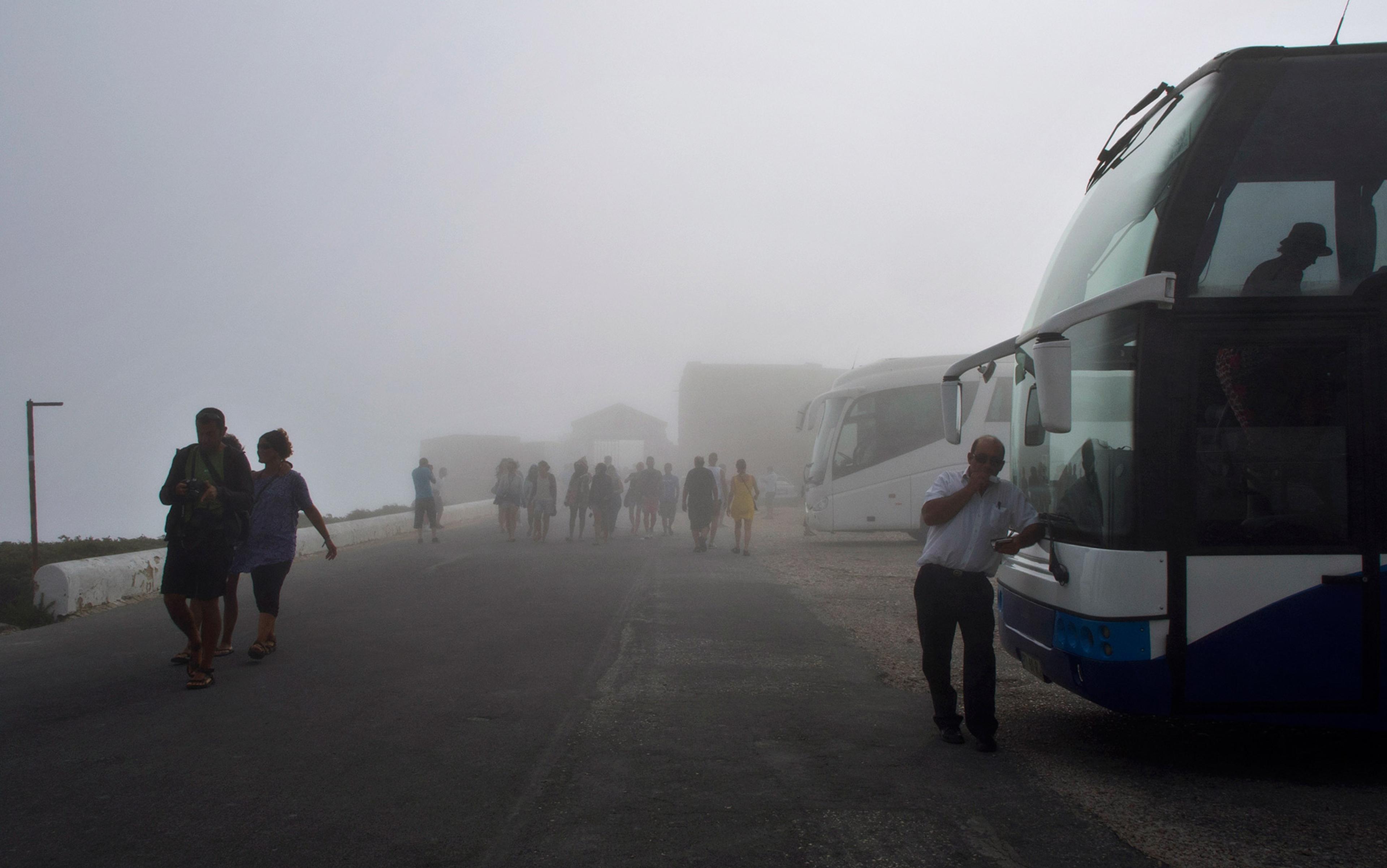 Tourists walking on a foggy road with buses parked on the side and a driver standing beside a bus, partially obstructed by the fog.