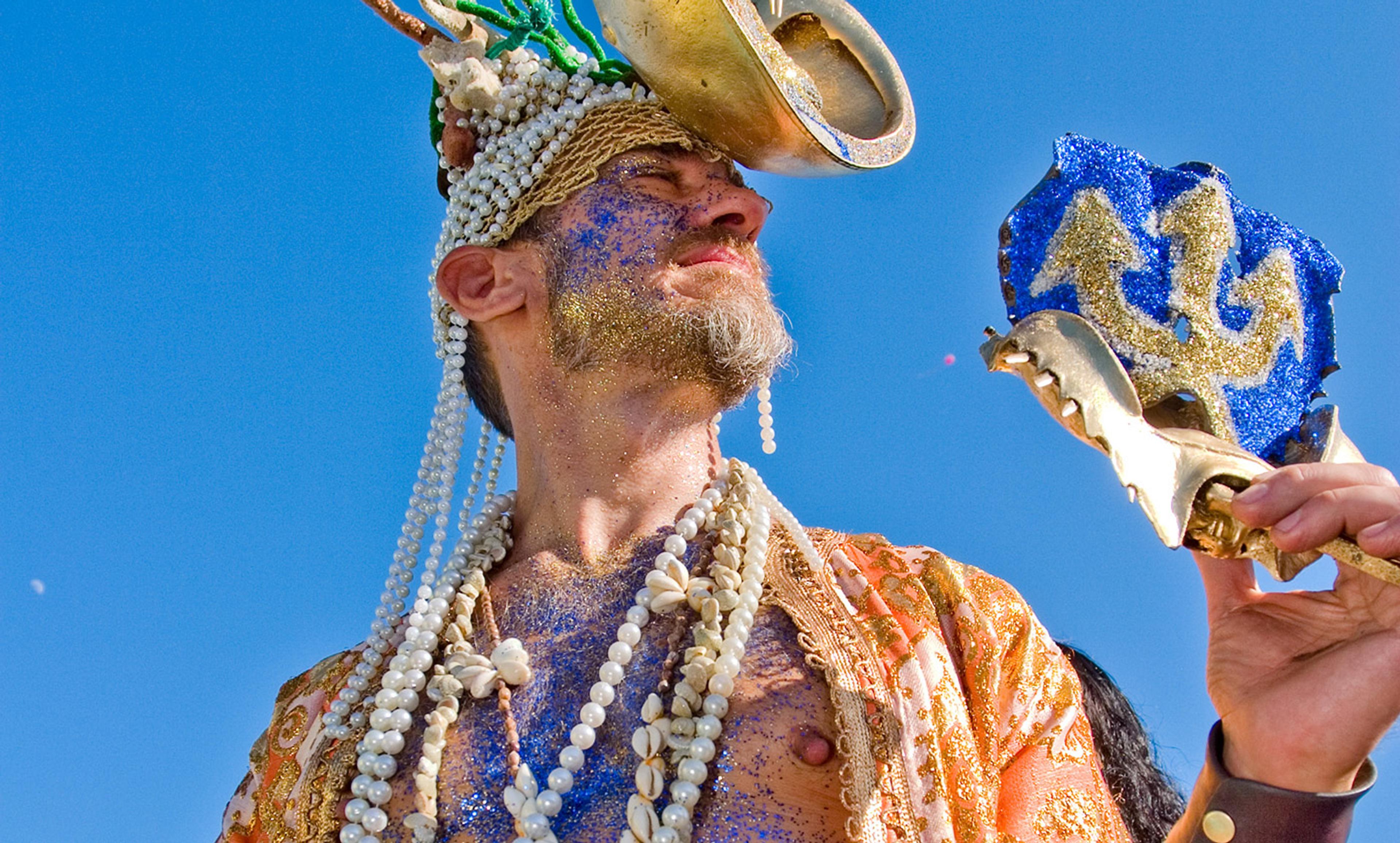 A person in colourful costume, adorned with shells and beads, holding a blue and gold trident prop against a clear blue sky.