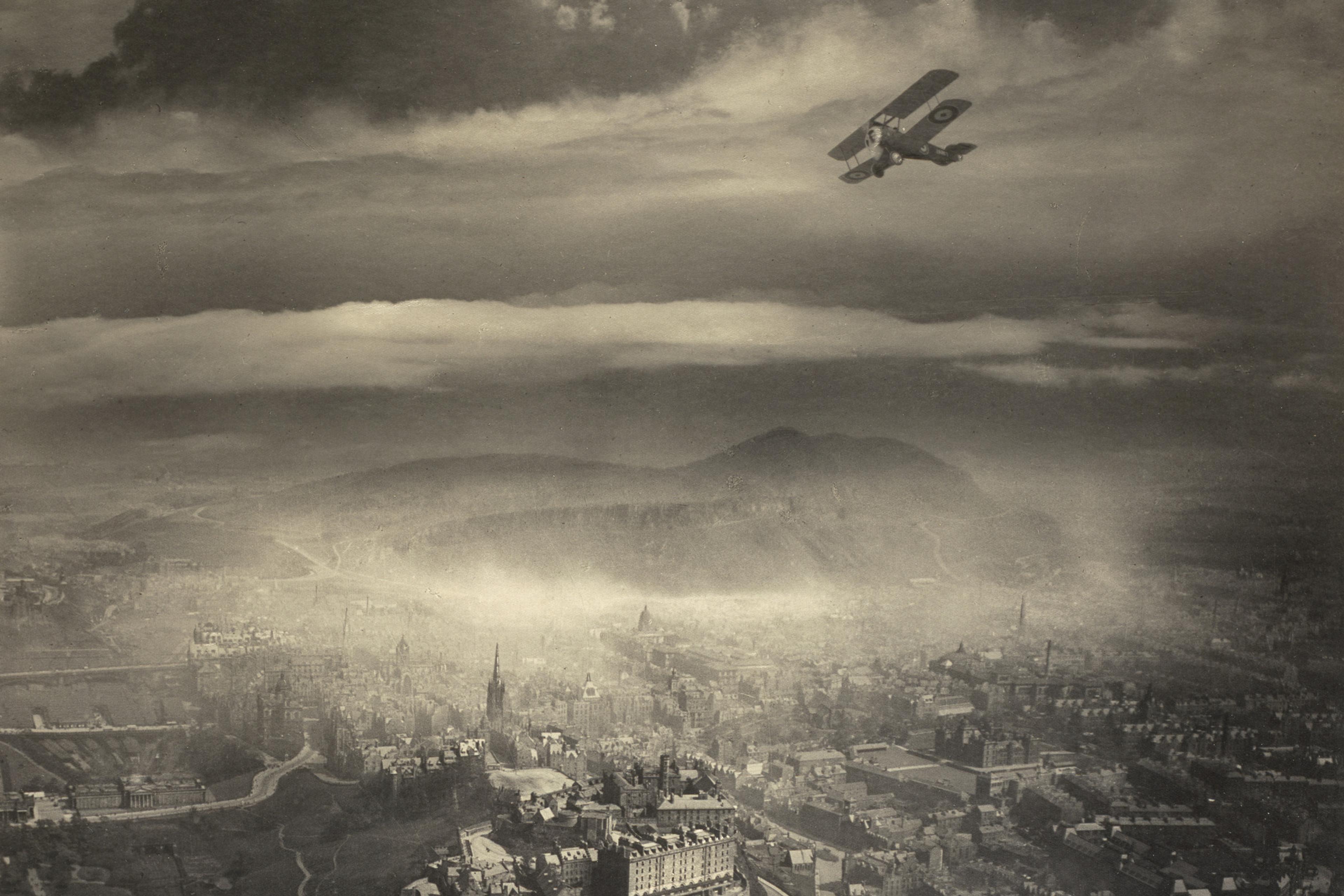 Aerial black and white photograph of a biplane flying over Edinburgh Castle and cityscape with dramatic clouds.