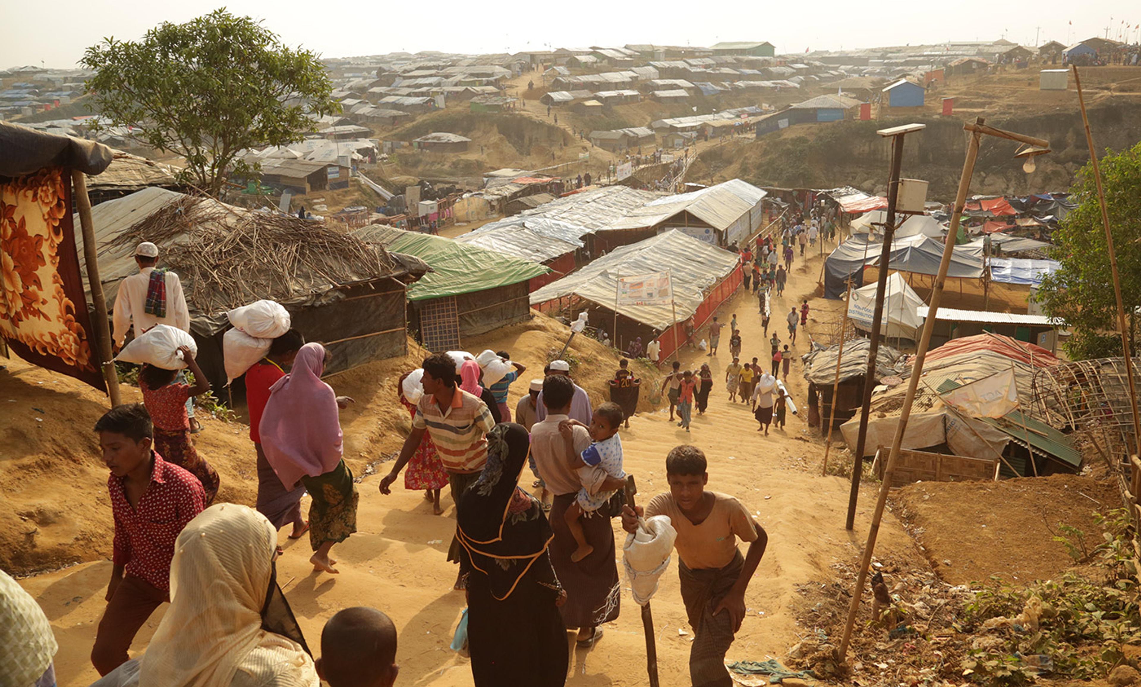 A refugee camp with numerous makeshift shelters and people walking along a dirt path, carrying supplies and children.
