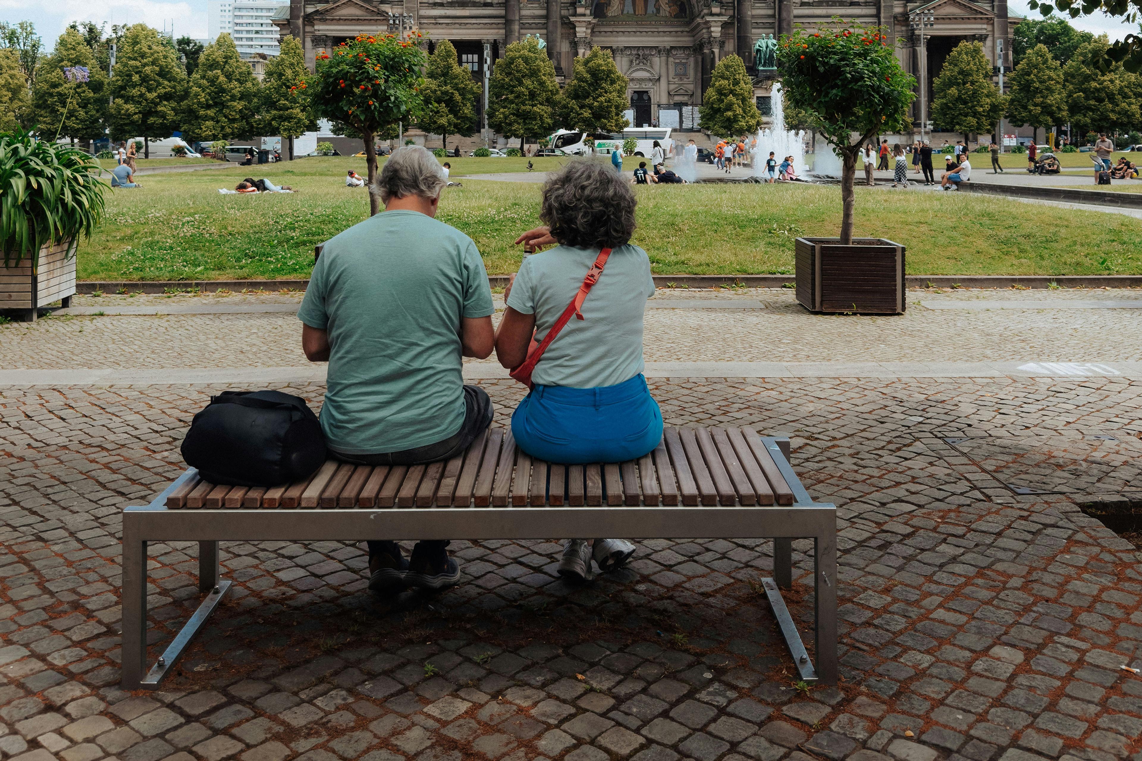An older couple sitting on a bench in a park with a historical building and fountain in the background.