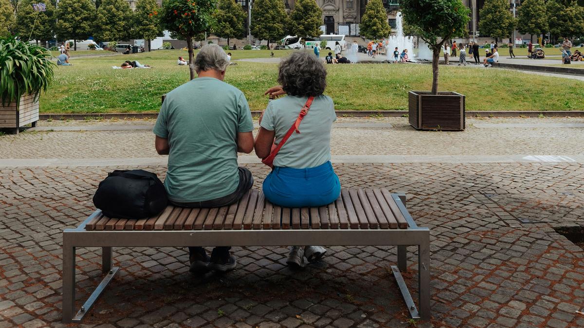 An older couple sitting on a bench in a park with a historical building and fountain in the background.