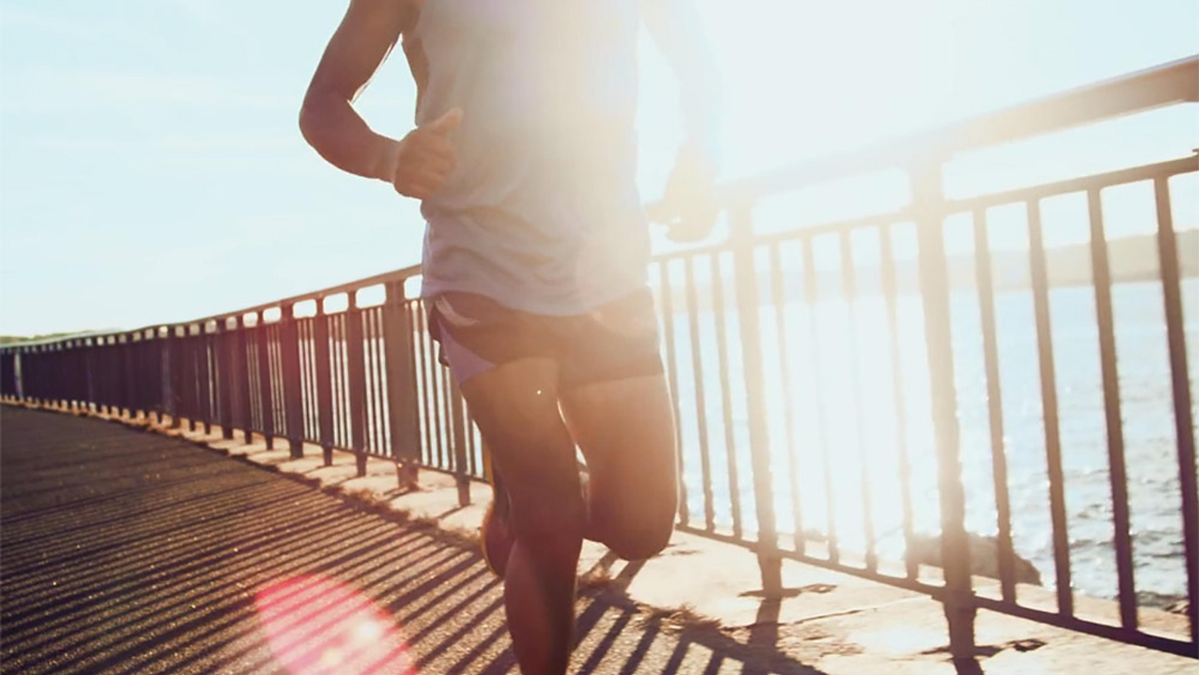 A person jogging on a waterfront path with sunlight shining through, creating lens flare and shadows on the ground.