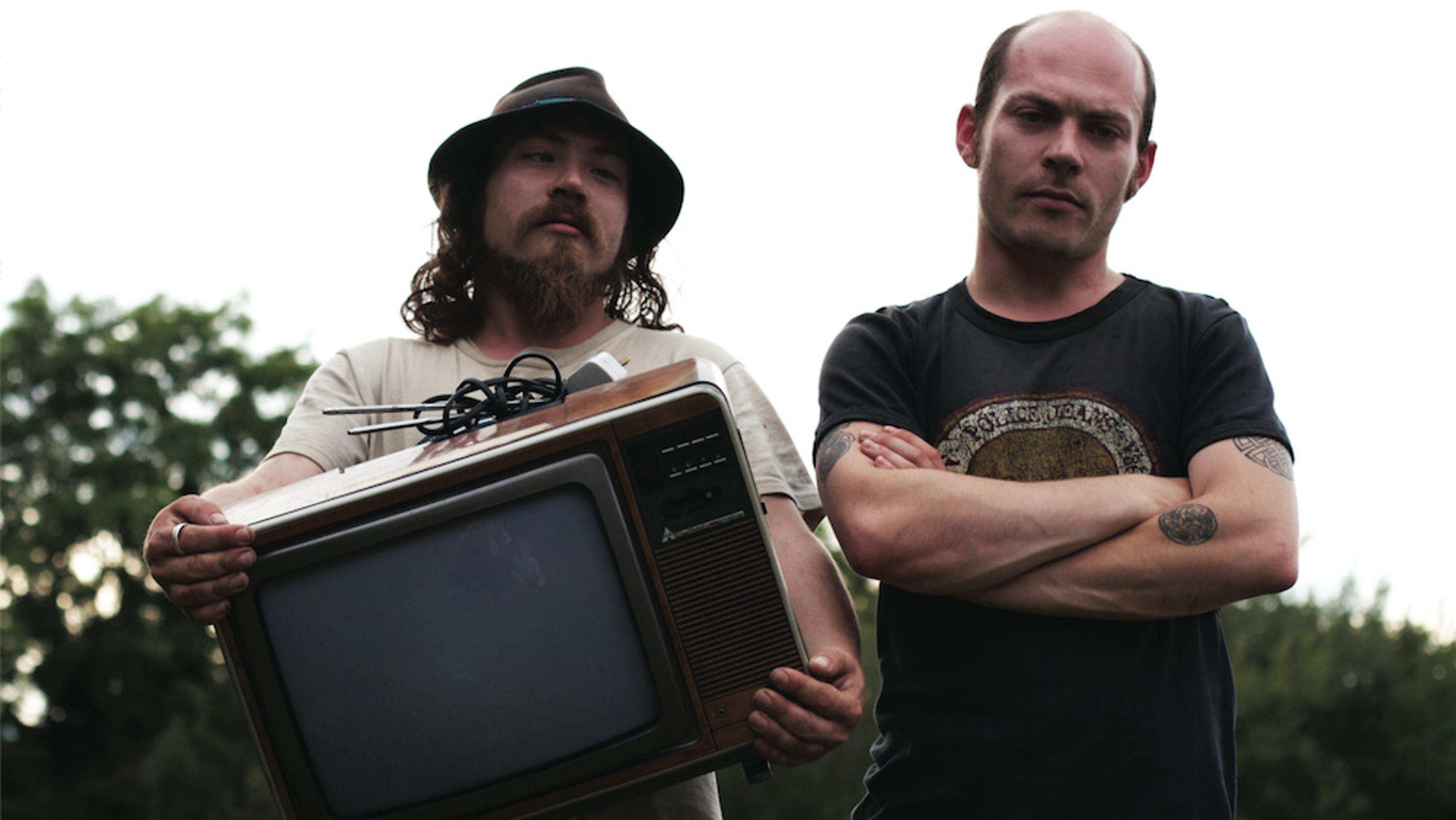 Two men outdoors, one with a hat holding an old television, the other crossing his arms with a tattoo on his forearm.