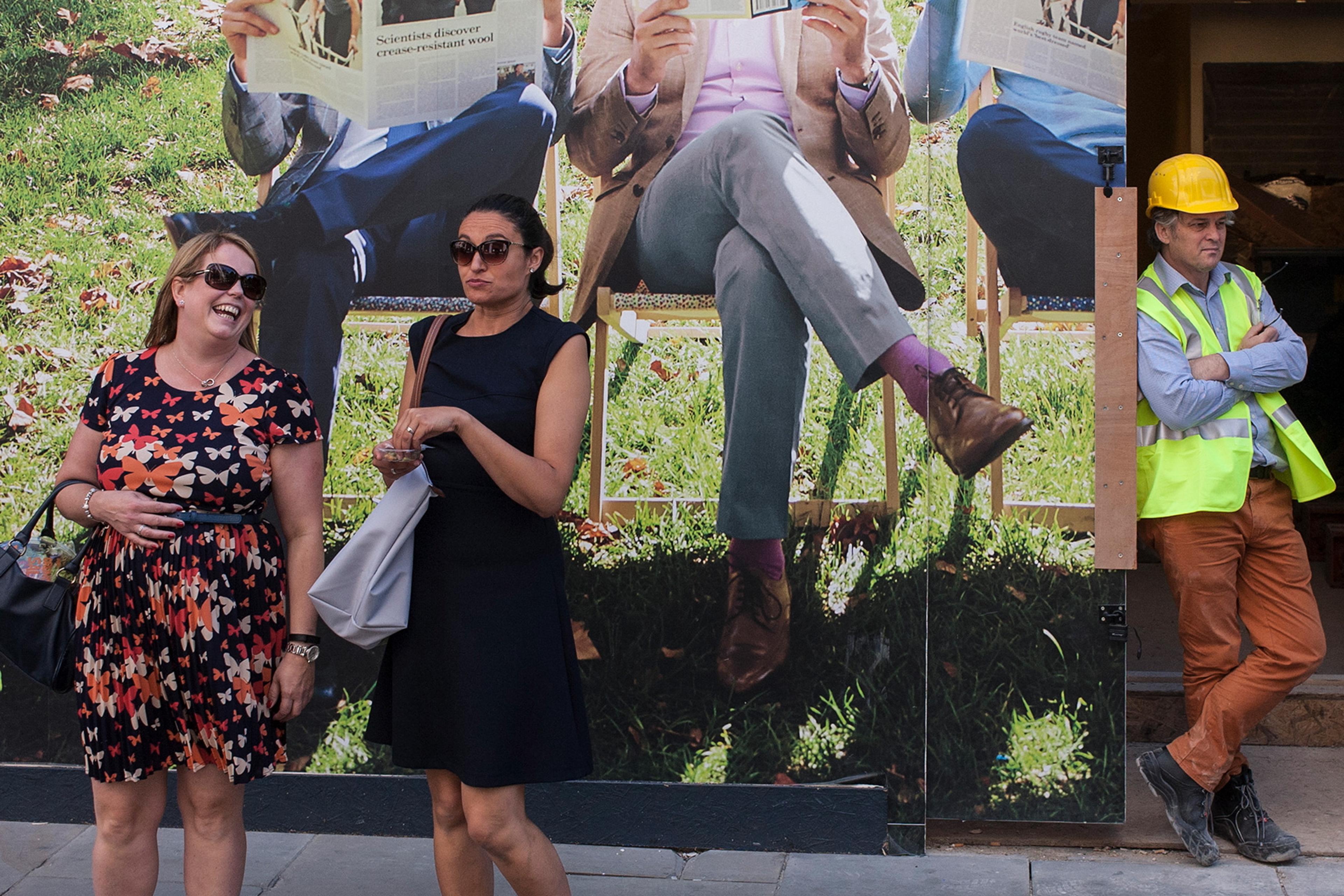 Photo of two women in front of a wall mural showing men reading newspapers, with a construction worker nearby.