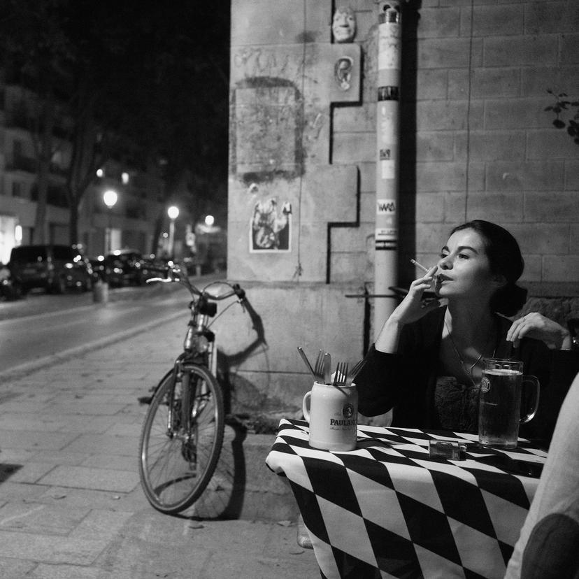 A woman sits at a cafe at night with a beer and a cigarette