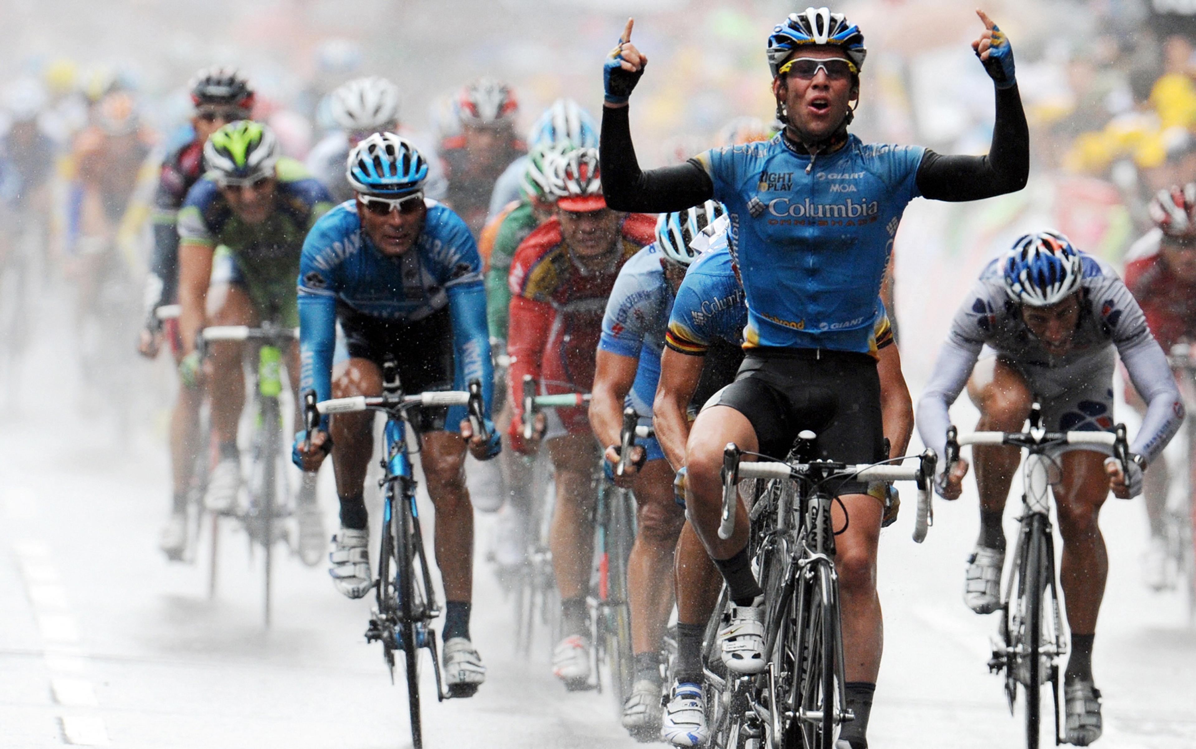 Cyclists in a professional race riding in rainy conditions. The leading cyclist in a blue jersey raises his arms in victory, with other cyclists closely following behind on the wet road. Everyone is wearing helmets and sunglasses for protection.