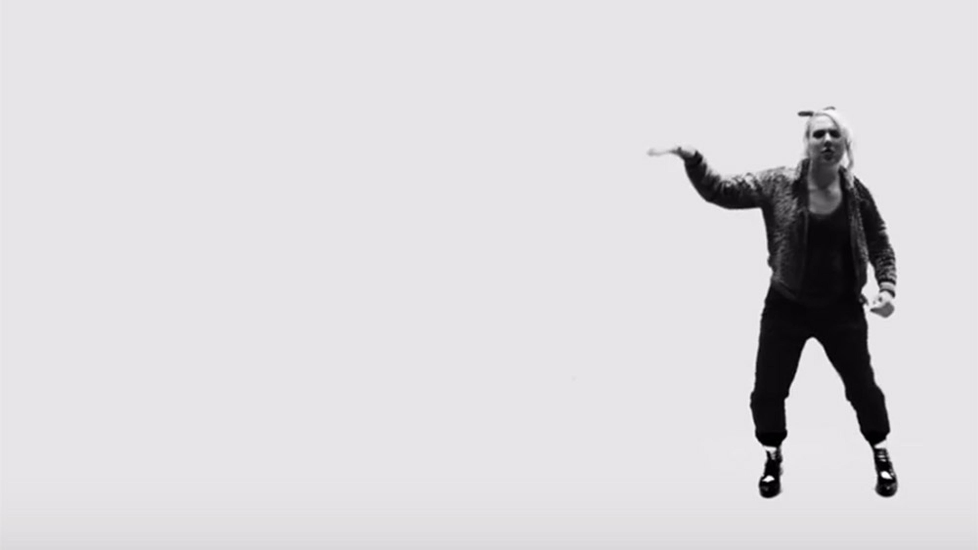 Black-and-white photo of a woman in casual clothes and boots posing, mid-movement, against a plain backdrop.