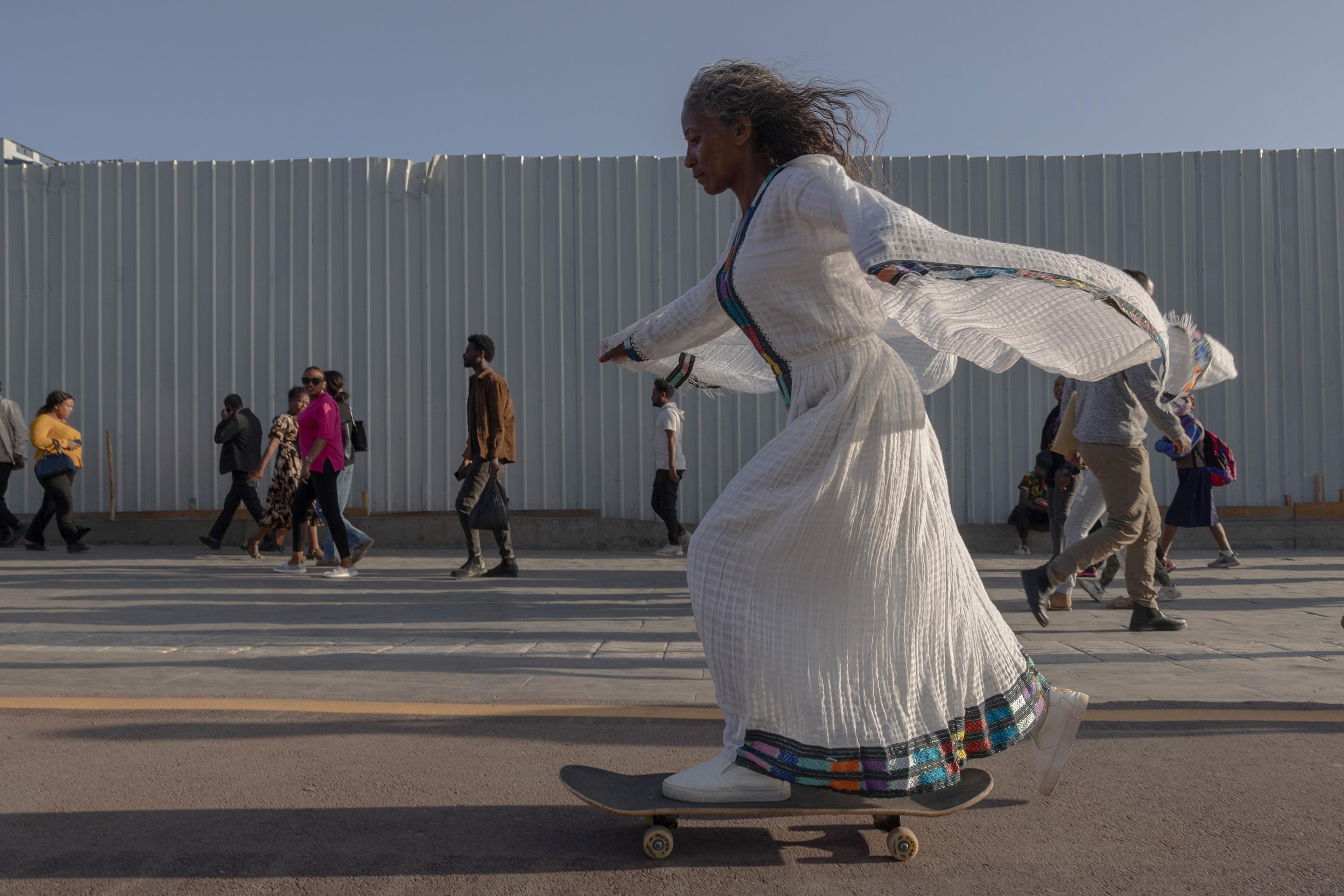 A woman in a flowing white dress skateboarding past people walking on a sunny day next to a corrugated fence.