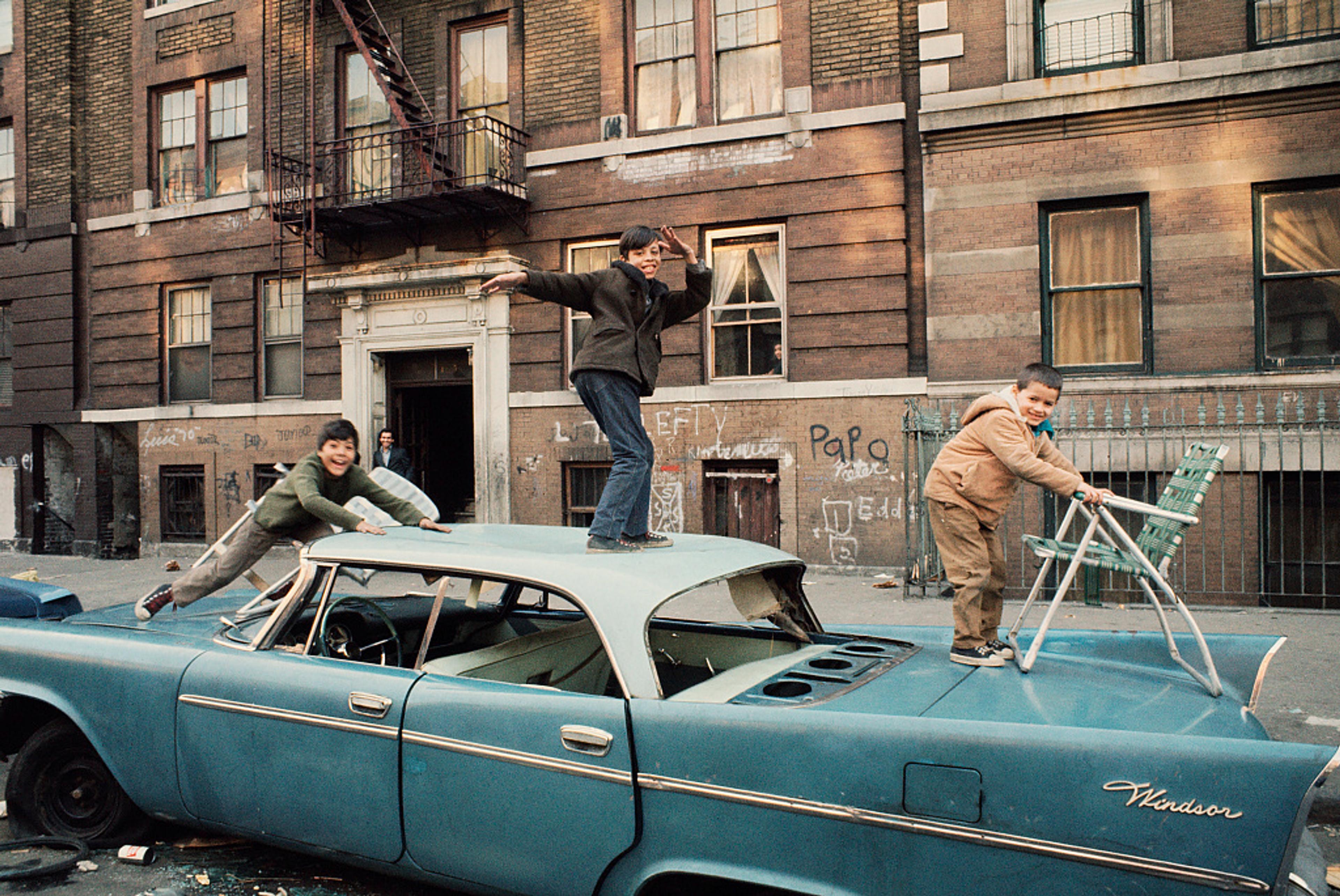Boys playing on the roof and hood of an abandoned 1950s car in an urban street covered in graffiti