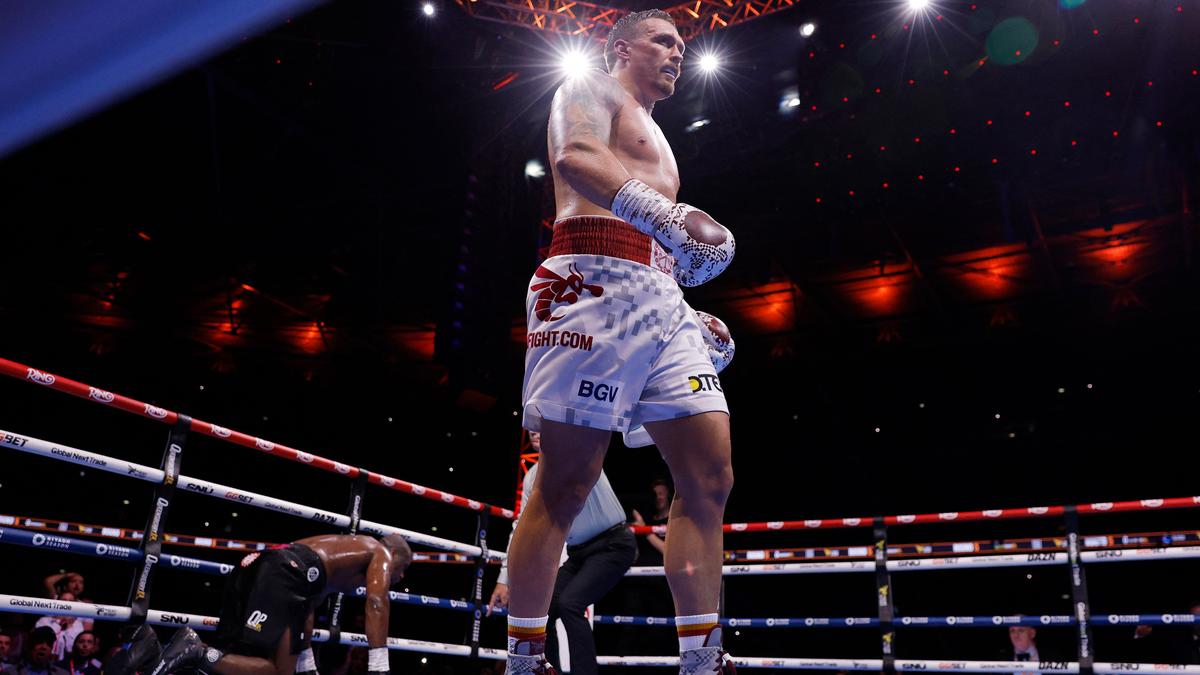 Photo of a boxing match with a standing boxer in the ring and an opponent kneeling behind, lit by overhead lights.