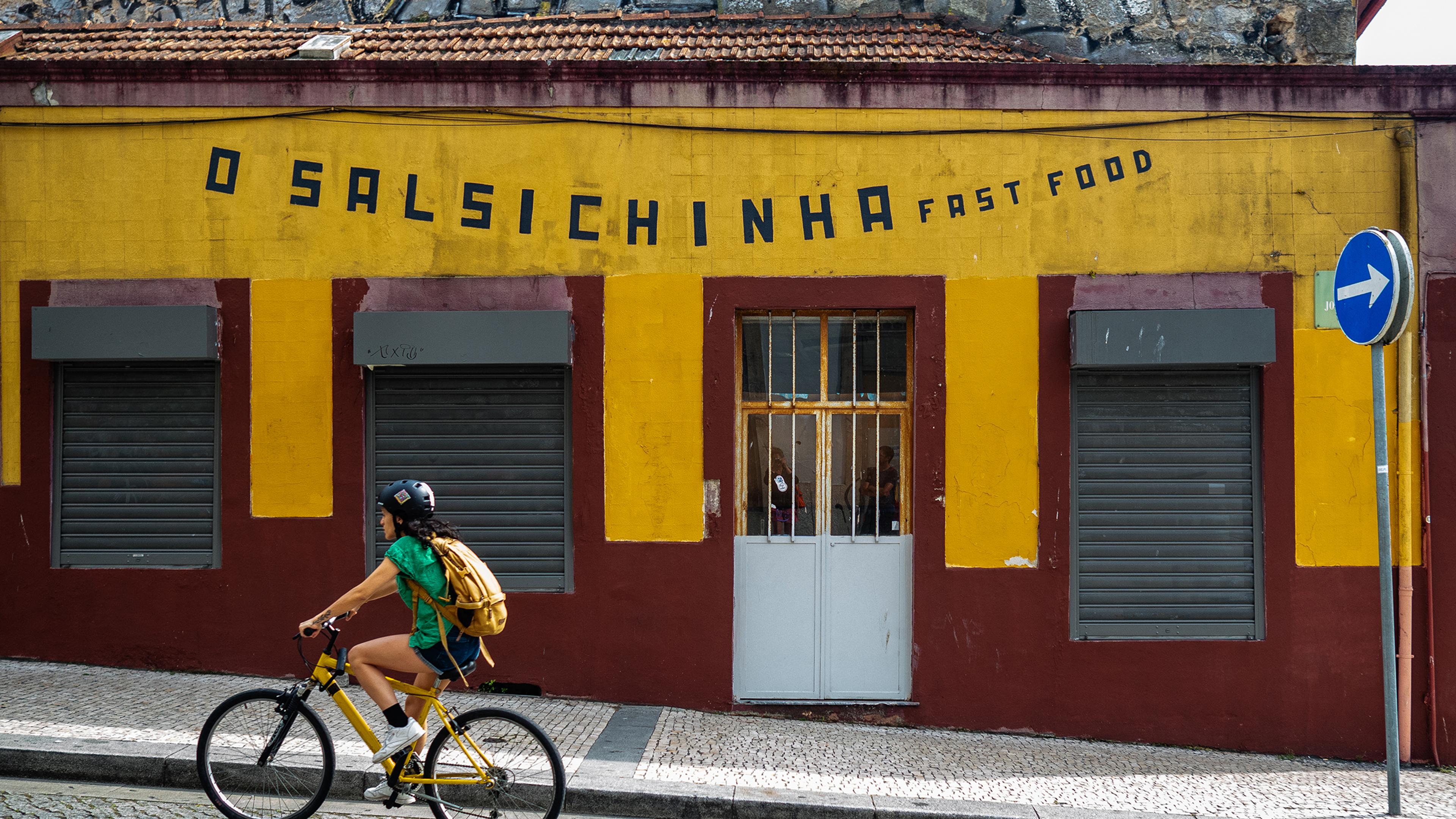 A cyclist on a yellow bike passing a closed fast food shop with a yellow and maroon facade and a one-way sign.