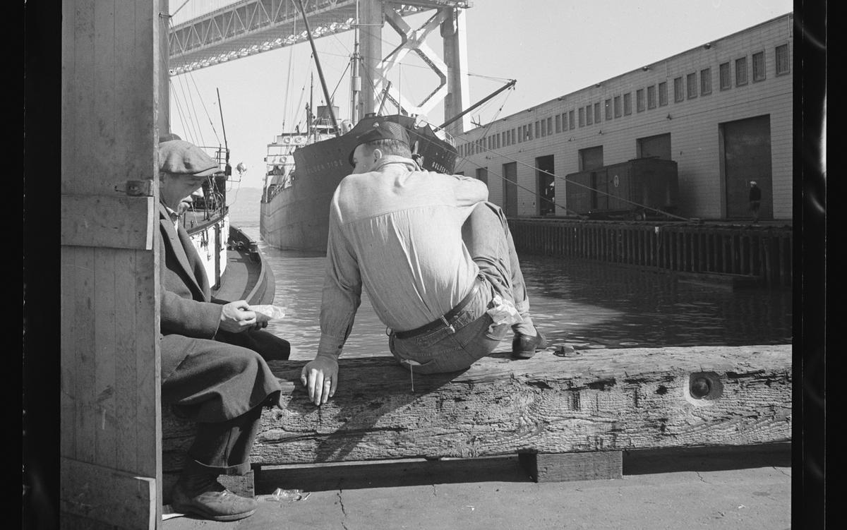 Two men sit talking on a wooden beam by a dock. One is in jeans, the other wears a coat and holds a small item. A cargo ship, a bridge and a warehouse are in the background.