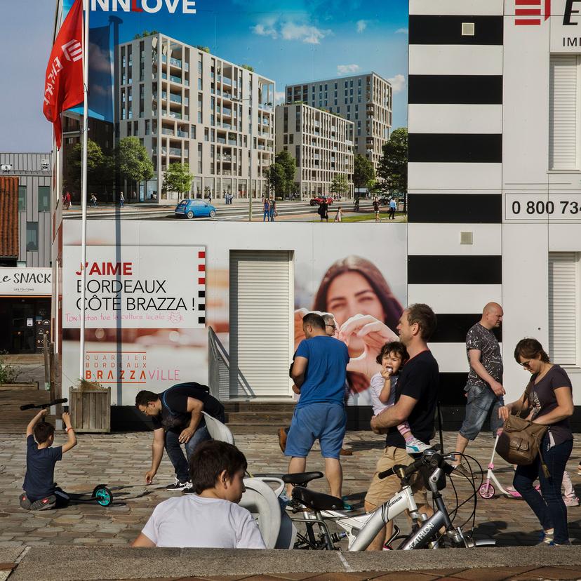 People sitting and walking through a city square on a sunny day, with a red brick building and large advertising posters covering other buildings.