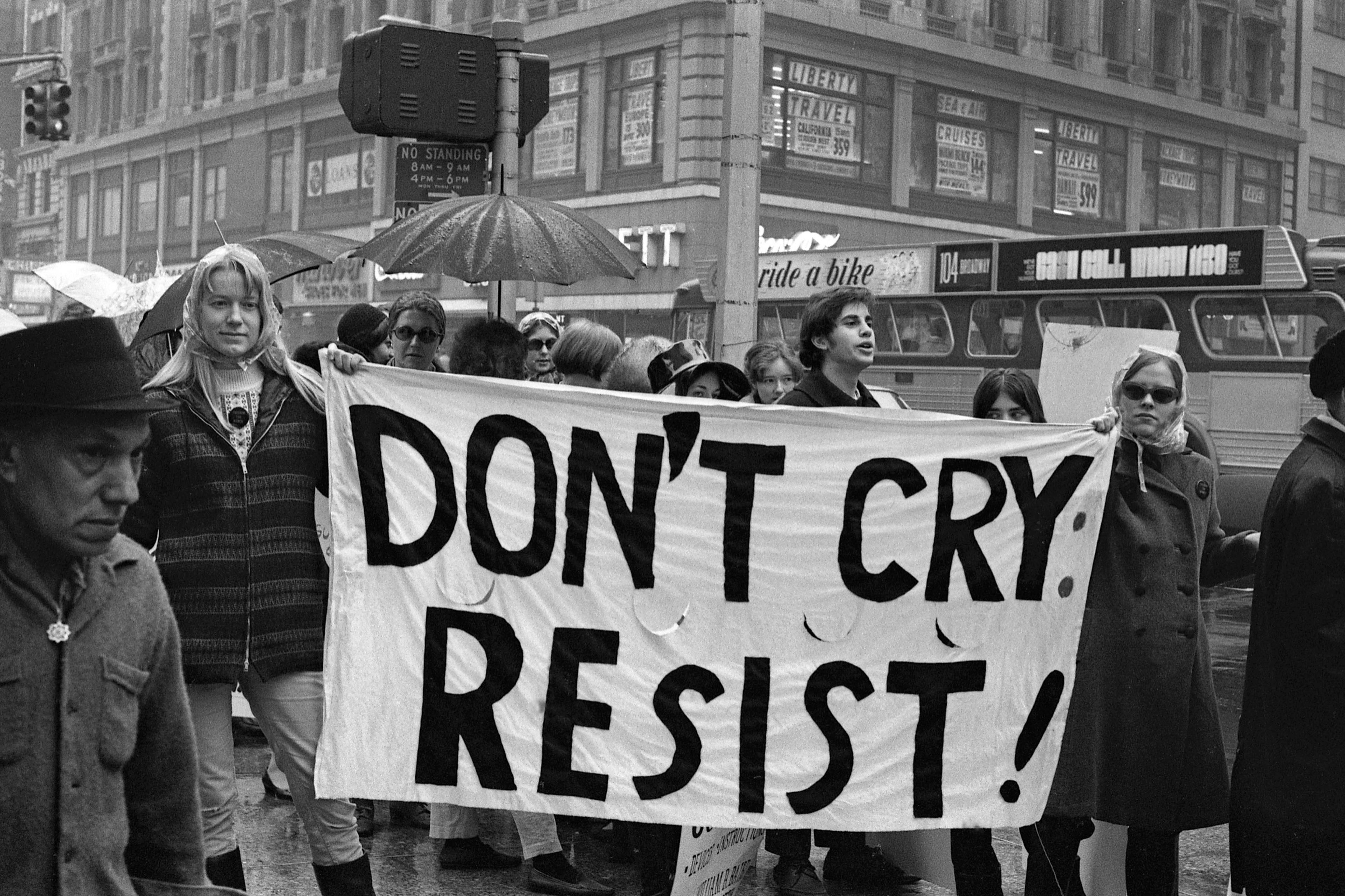 Black and white photo of protesters holding a banner saying “DON’T CRY: RESIST!” on a city street in the rain.
