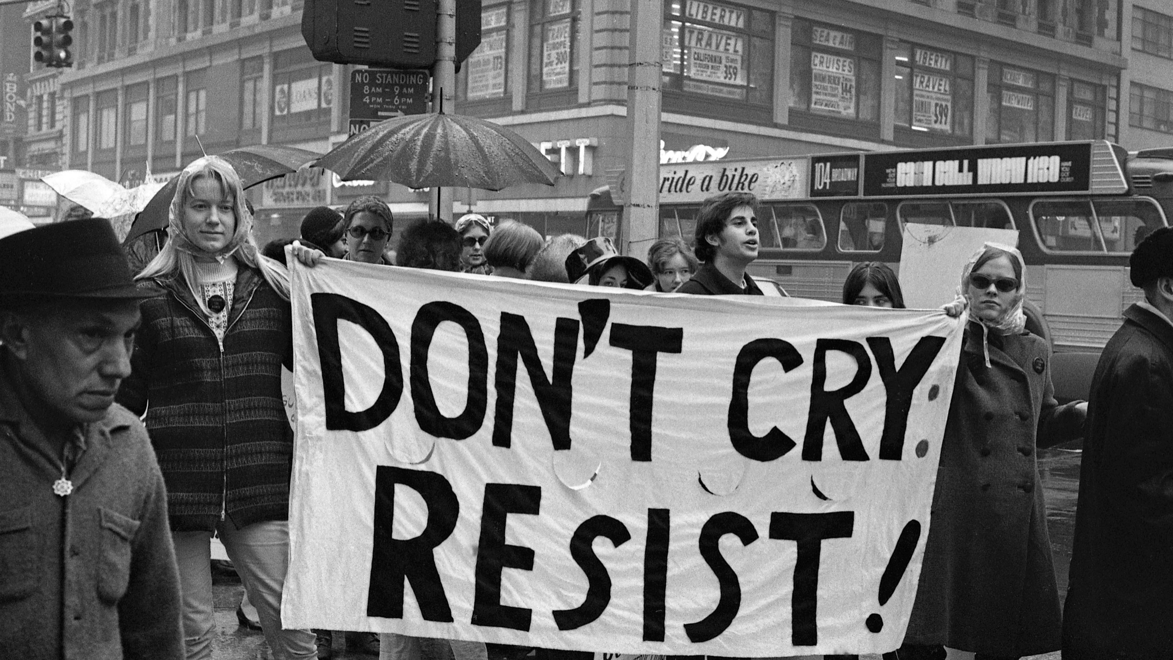 Black and white photo of protesters holding a banner saying “DON’T CRY: RESIST!” on a city street in the rain.