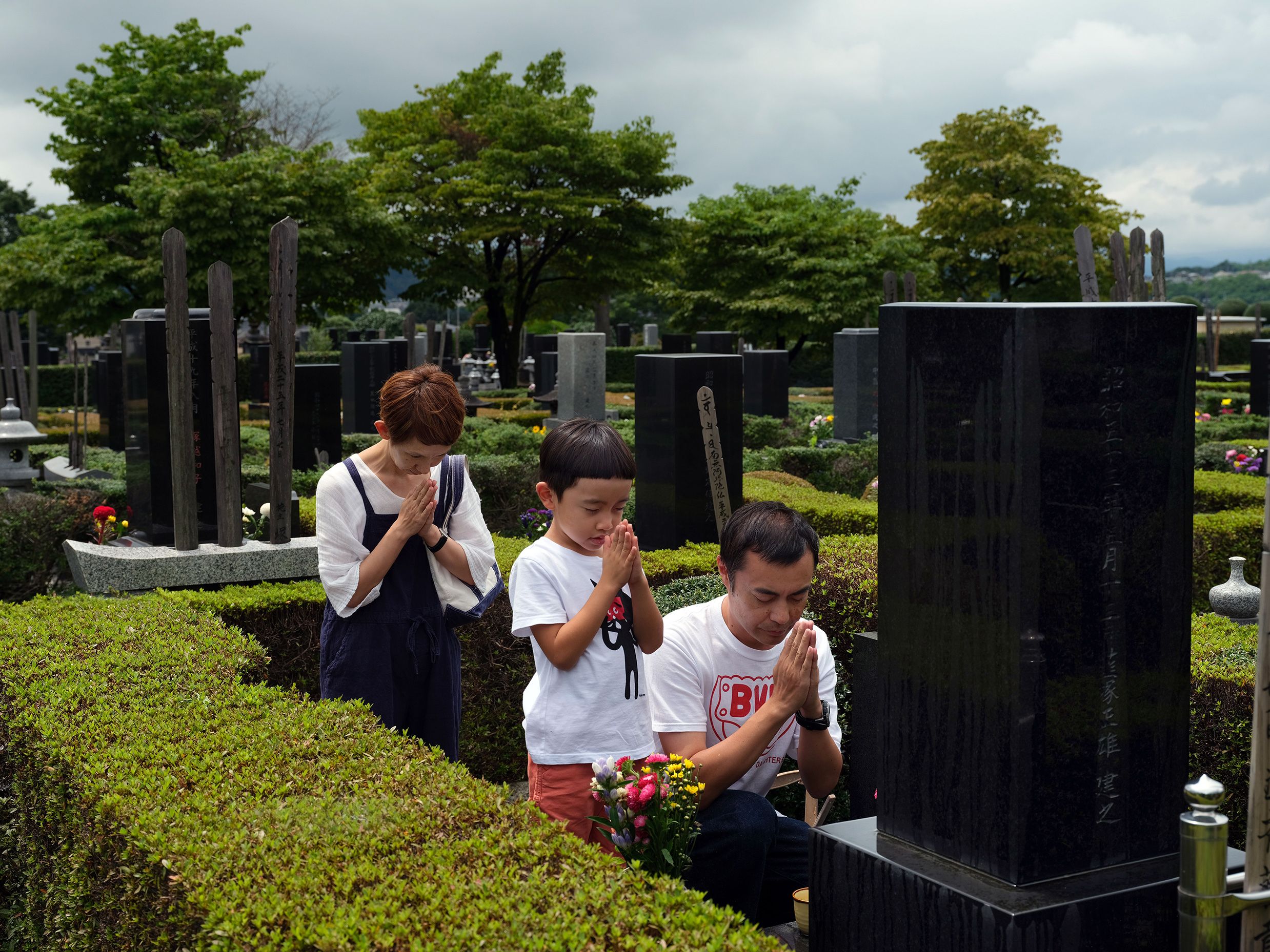 A family praying at a cemetery, standing beside a grave with trees and tombstones in the background.