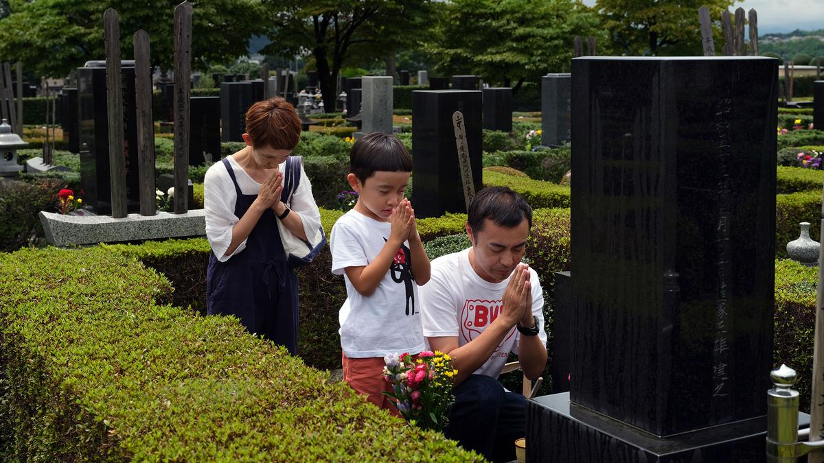 A family praying at a cemetery, standing beside a grave with trees and tombstones in the background.