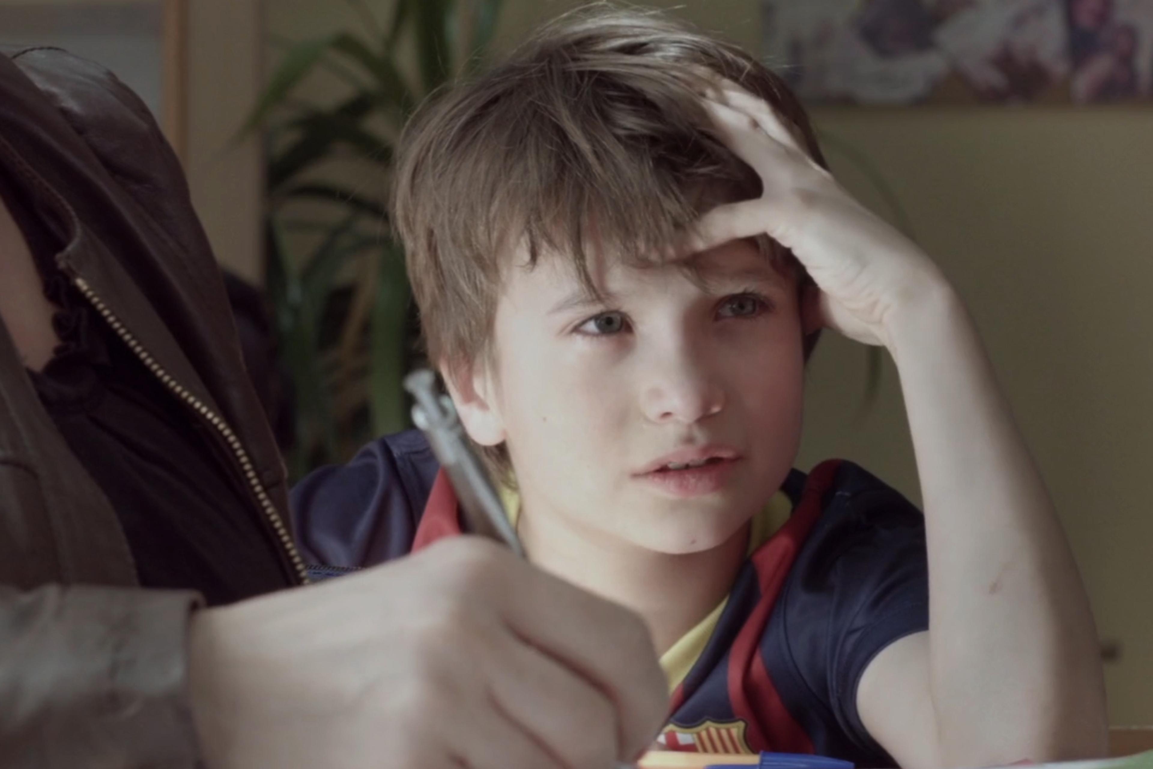 A young boy with a thoughtful expression sits at a table, holding a pen, with one hand on his head. A person next to him is partially visible.