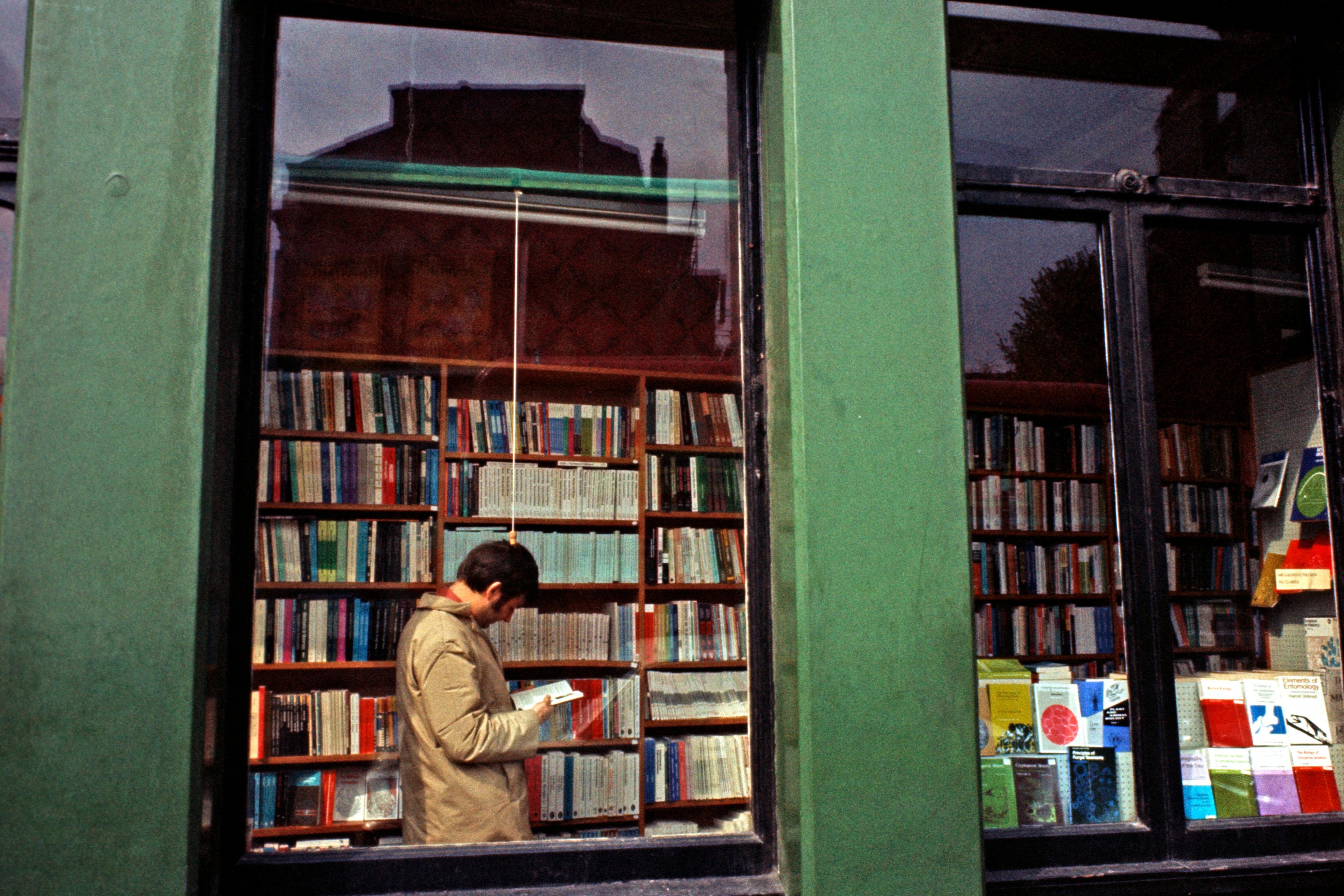 Photo of a bookshop exterior with green columns and a window displaying books. A person reads inside.