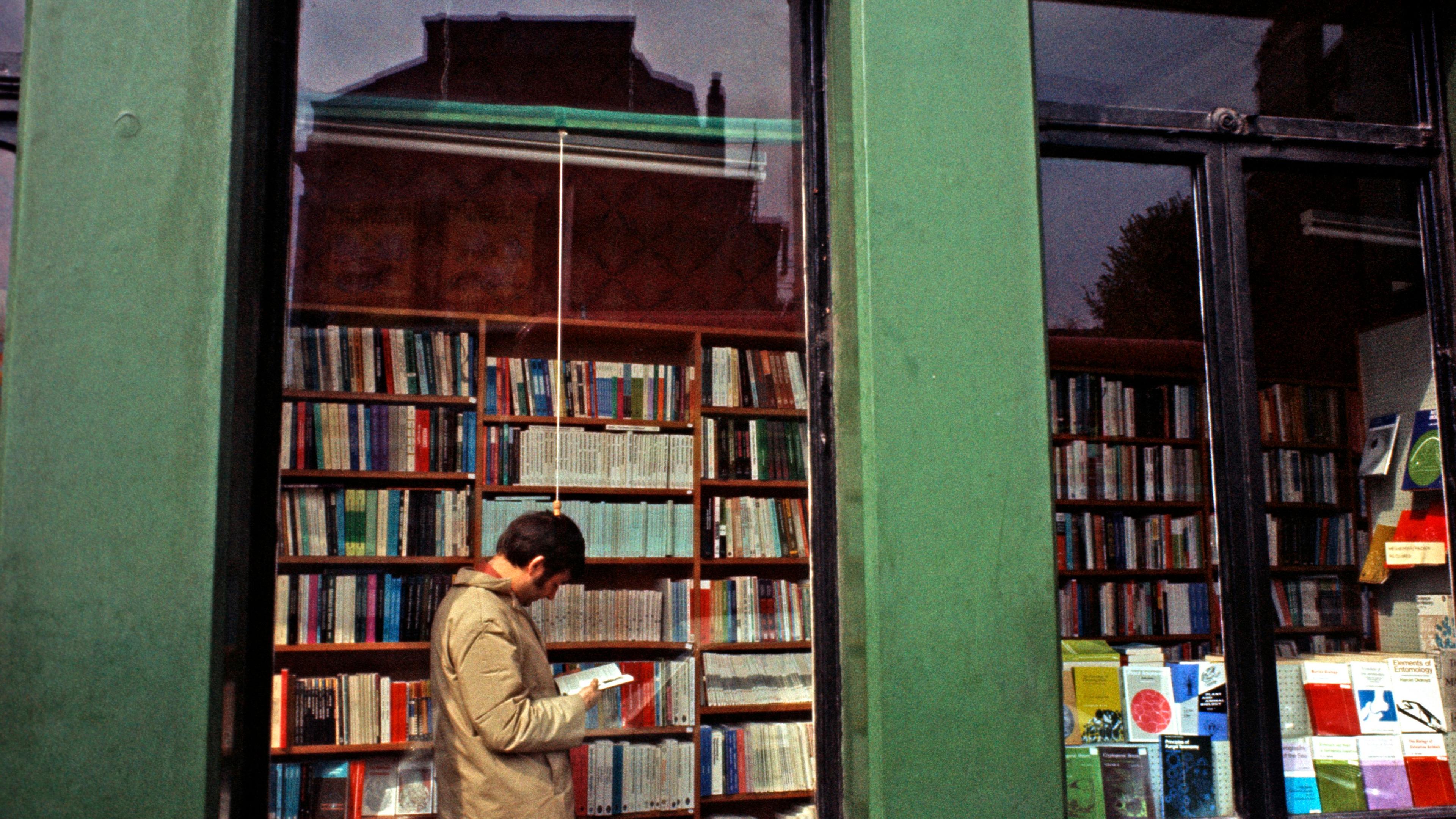 Photo of a bookshop exterior with green columns and a window displaying books. A person reads inside.