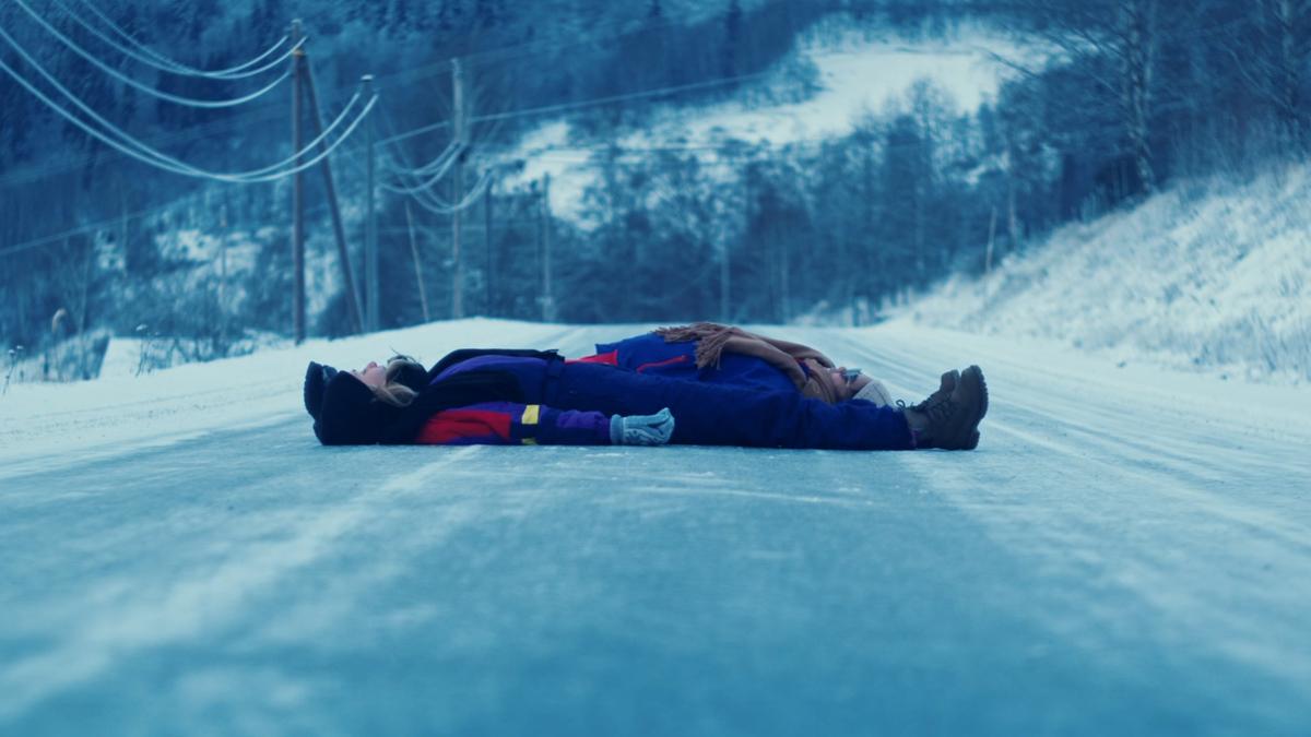 Two people lying on a snowy road in winter clothing with a forested landscape in the background.