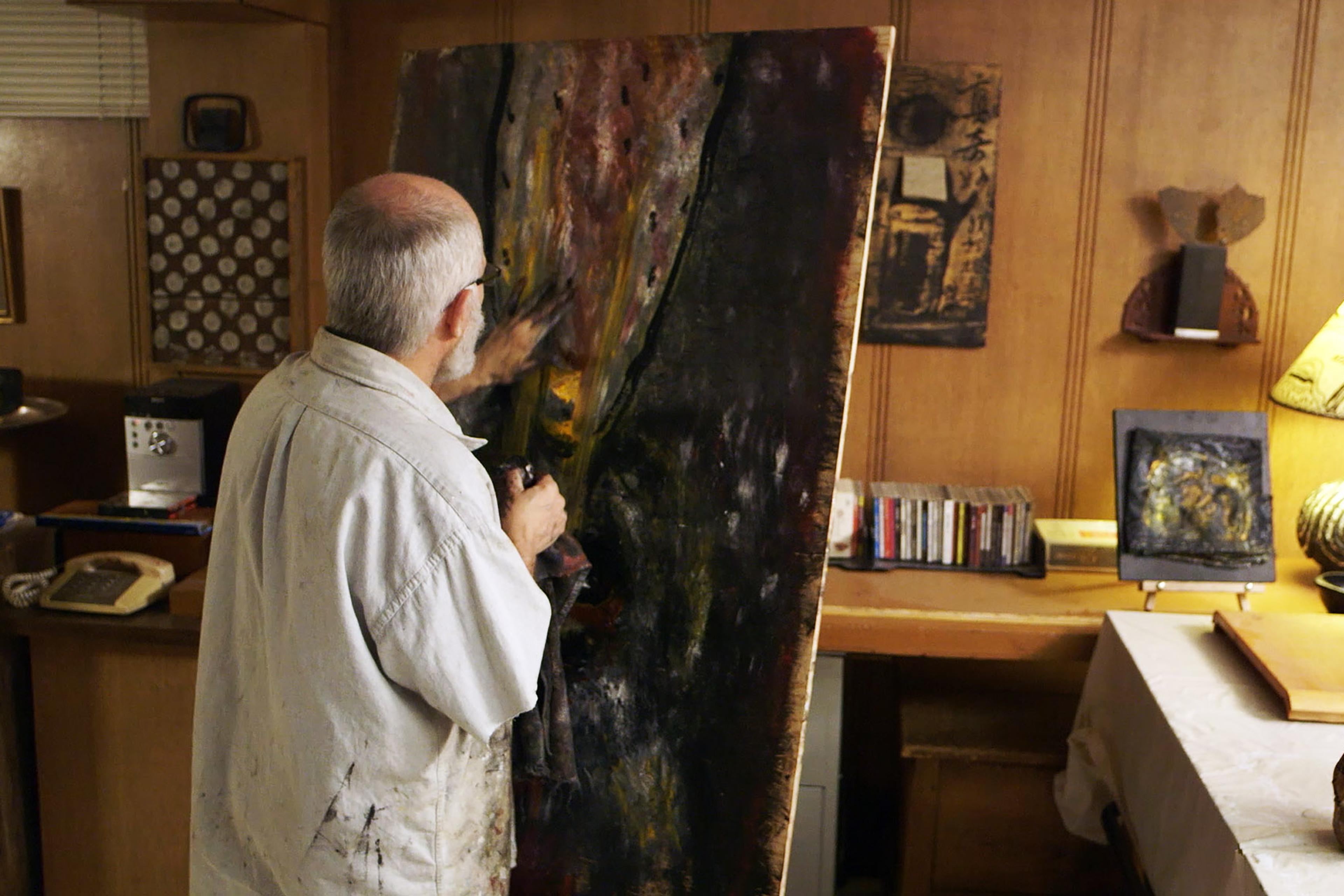 An older man creating abstract art on a large canvas in a cluttered room with artwork, books, and a lamp on a desk.