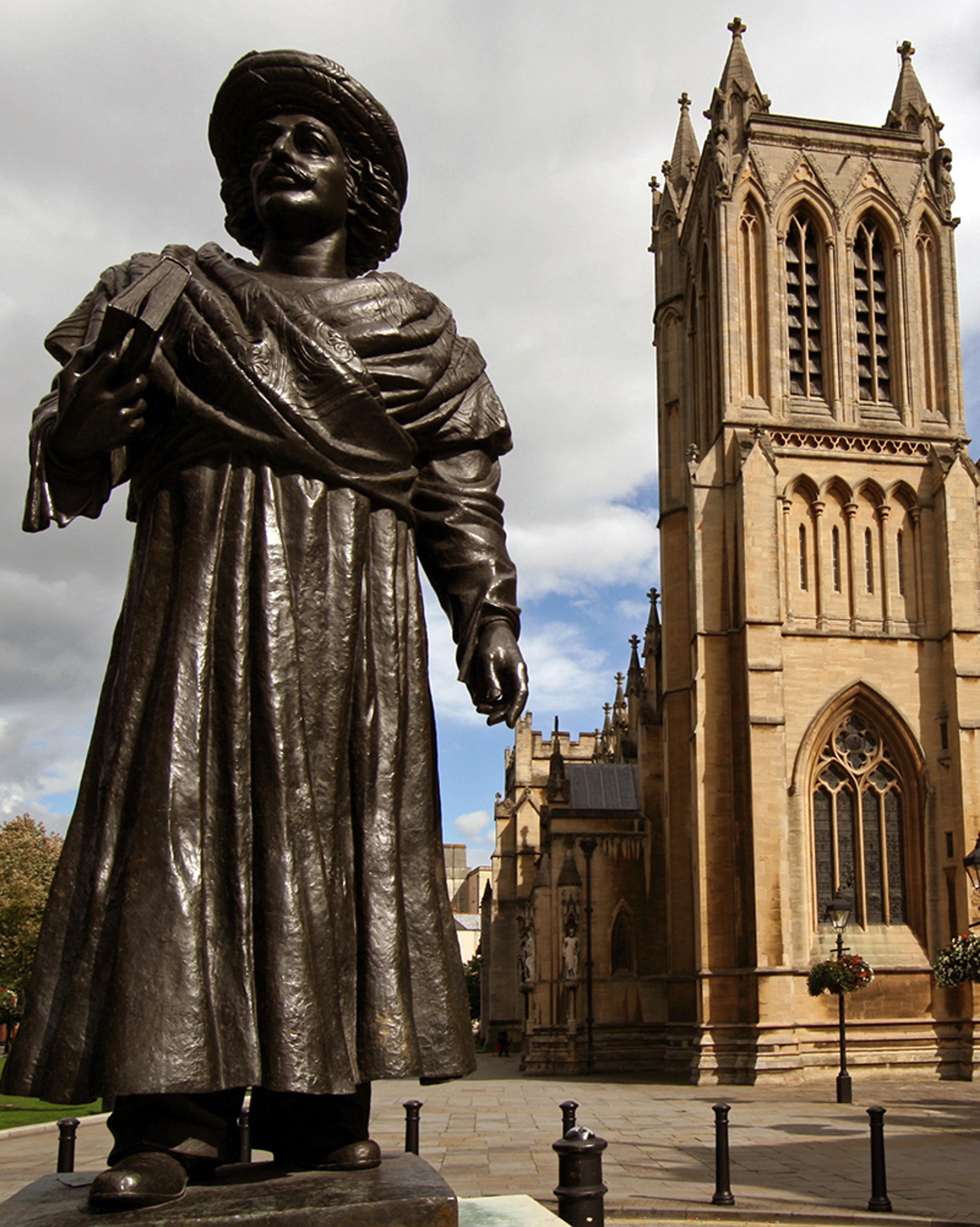 A bronze statue of a man in front of Bristol Cathedral with clouds and blue sky in the background.